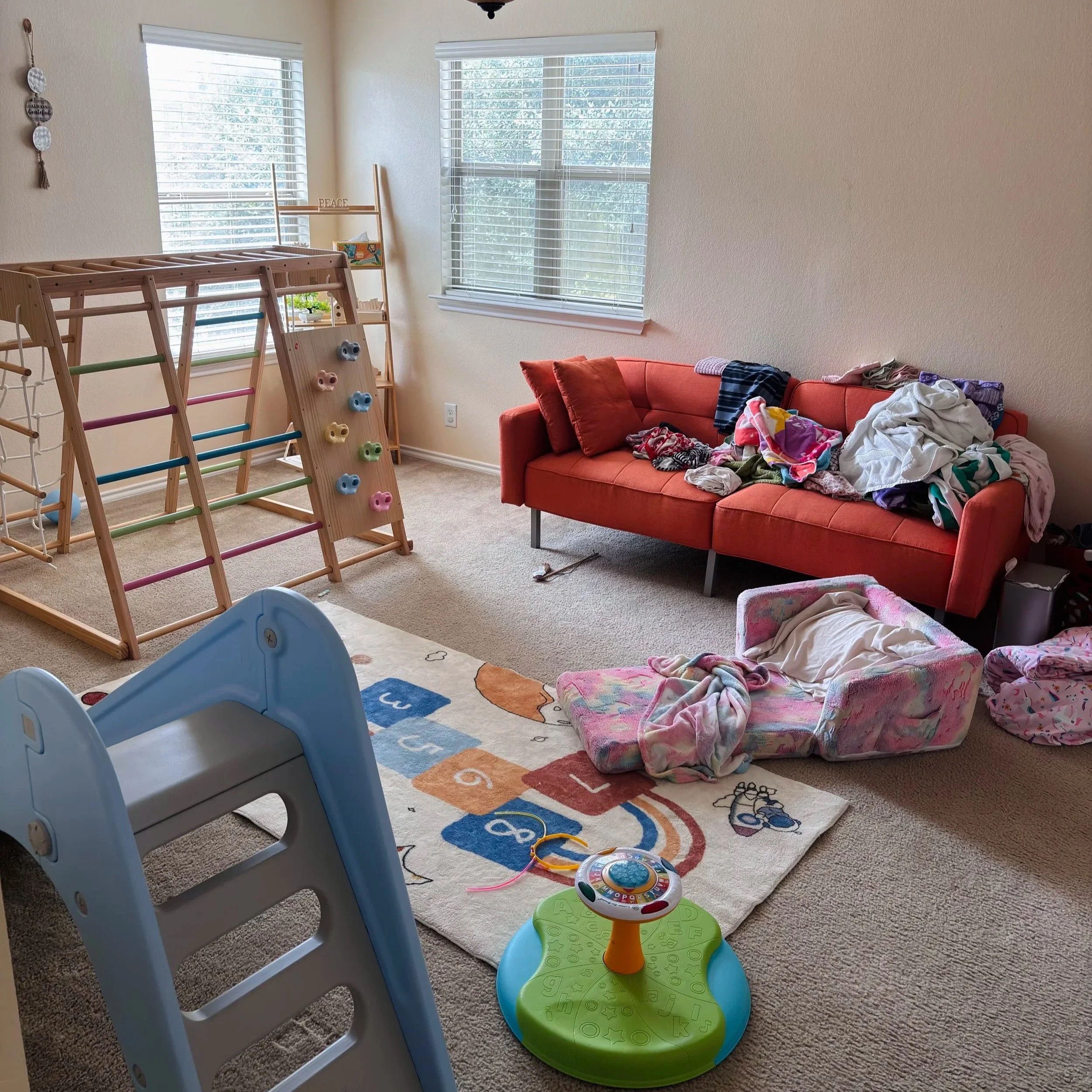 A cluttered living room with a red couch covered in blankets and clothes, a bright colorful baby activity center, a climbing wall with pastel-colored holds, a wooden step stool, and large windows with white blinds letting in natural light.