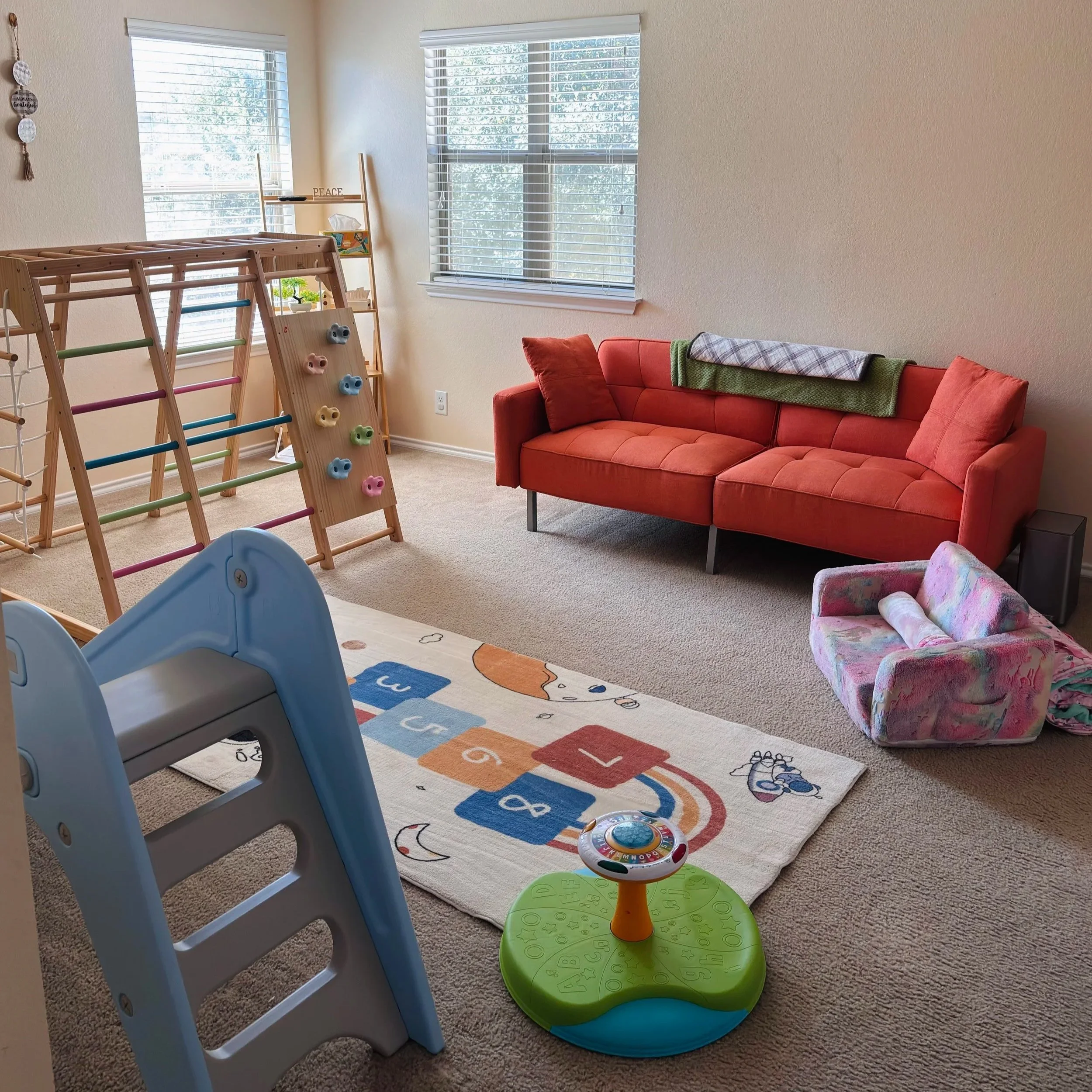 A playroom with a beige carpet, featuring a blue slide, a colorful climbing wall, a red couch with pillows and a blanket, a pink armchair, two large windows with white blinds, a small green table, and a rug with alphabet and number designs.
