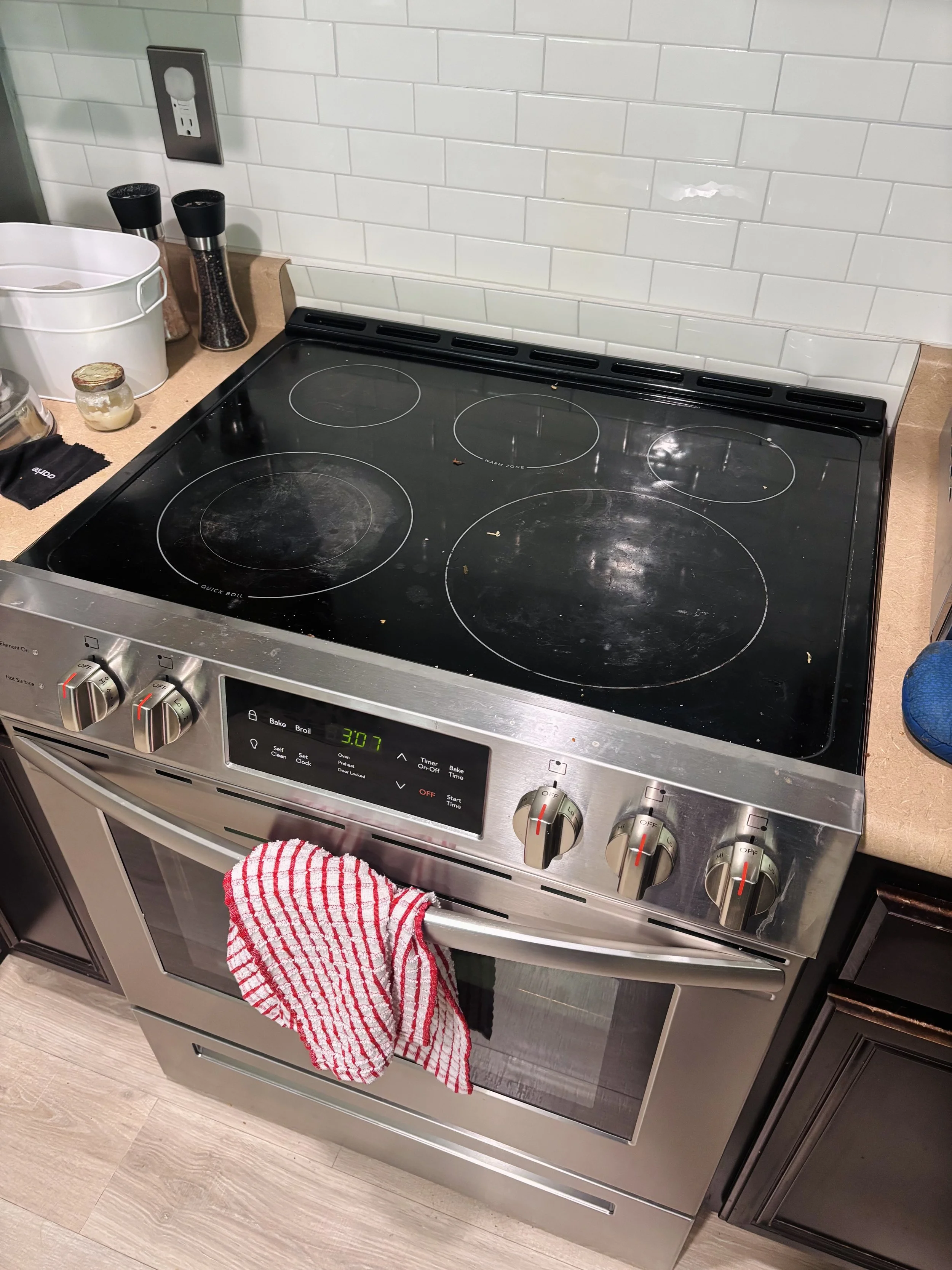 A stainless steel electric stove with a red and white checked kitchen towel hanging on its handle, set on a wooden floor with a white subway tile backsplash. The stove has four burners, control knobs, and a digital clock displaying 3:07.