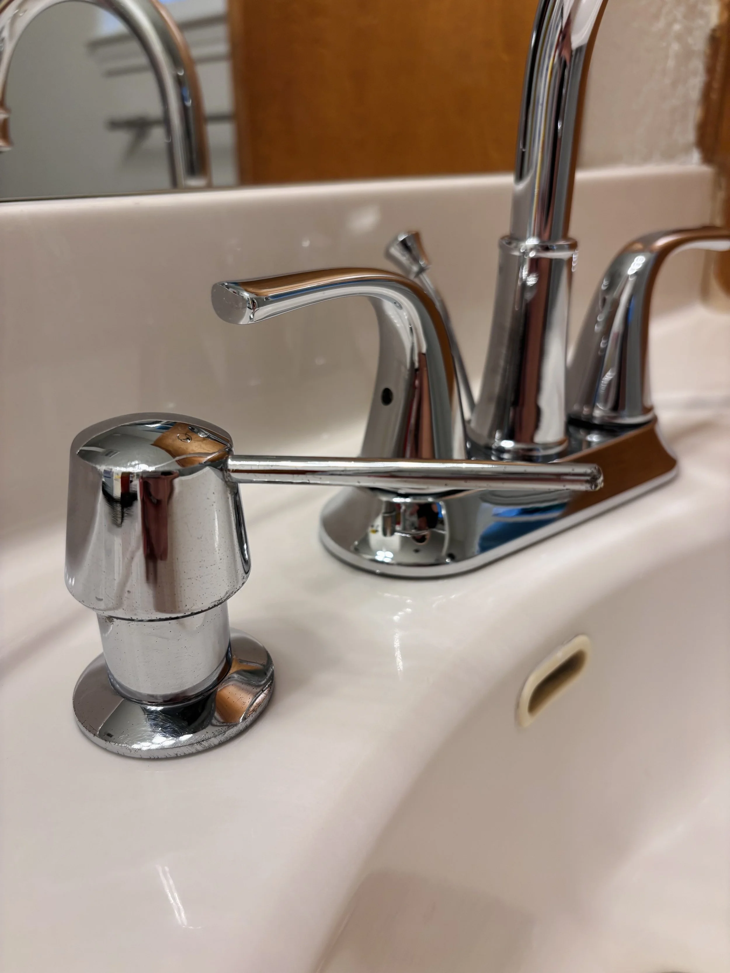 Close-up of a chrome bathroom faucet and handle on a white sink with a mirror and a towel rack in the background.