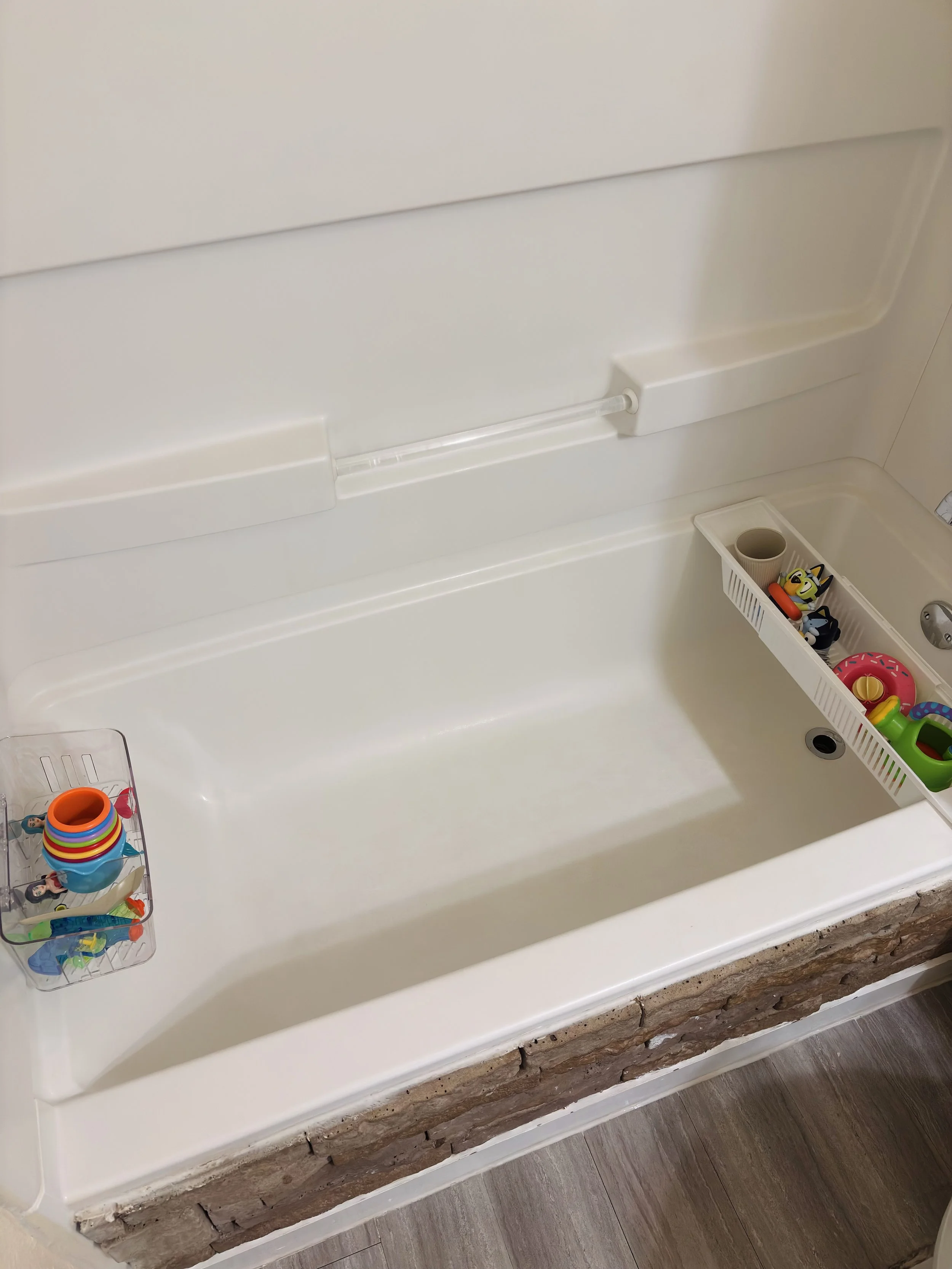The image shows the inside of a bathtub with a built-in shelf and a decorative stone border at the bottom front, with various colorful toys and a cup in a basket and on the shelf.