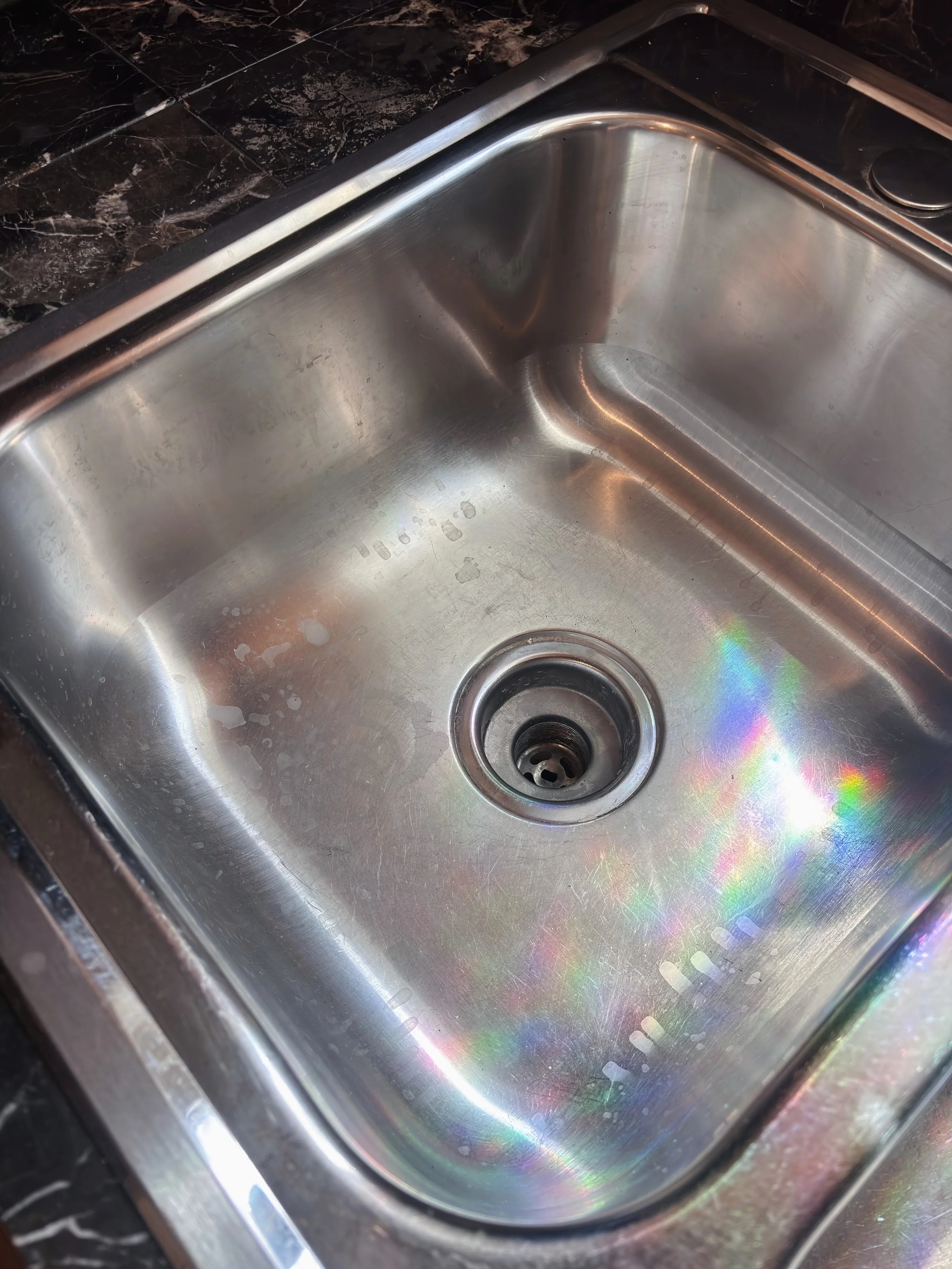 A stainless steel kitchen sink with some water stains and a small rainbow reflection near the drain.