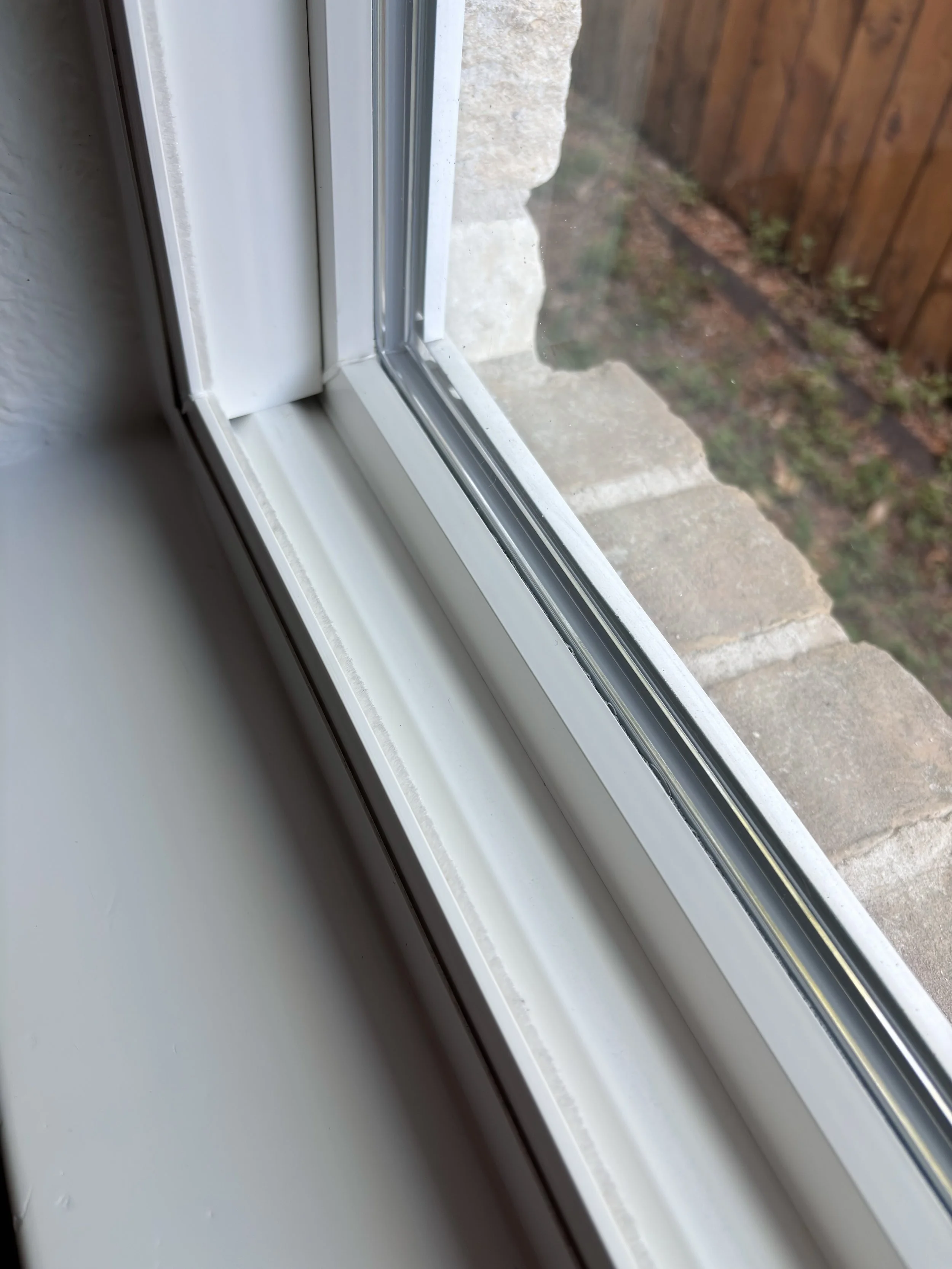 Close-up view of a white window frame and sliding window glass, with a view of a brick wall and a wooden fence outside.