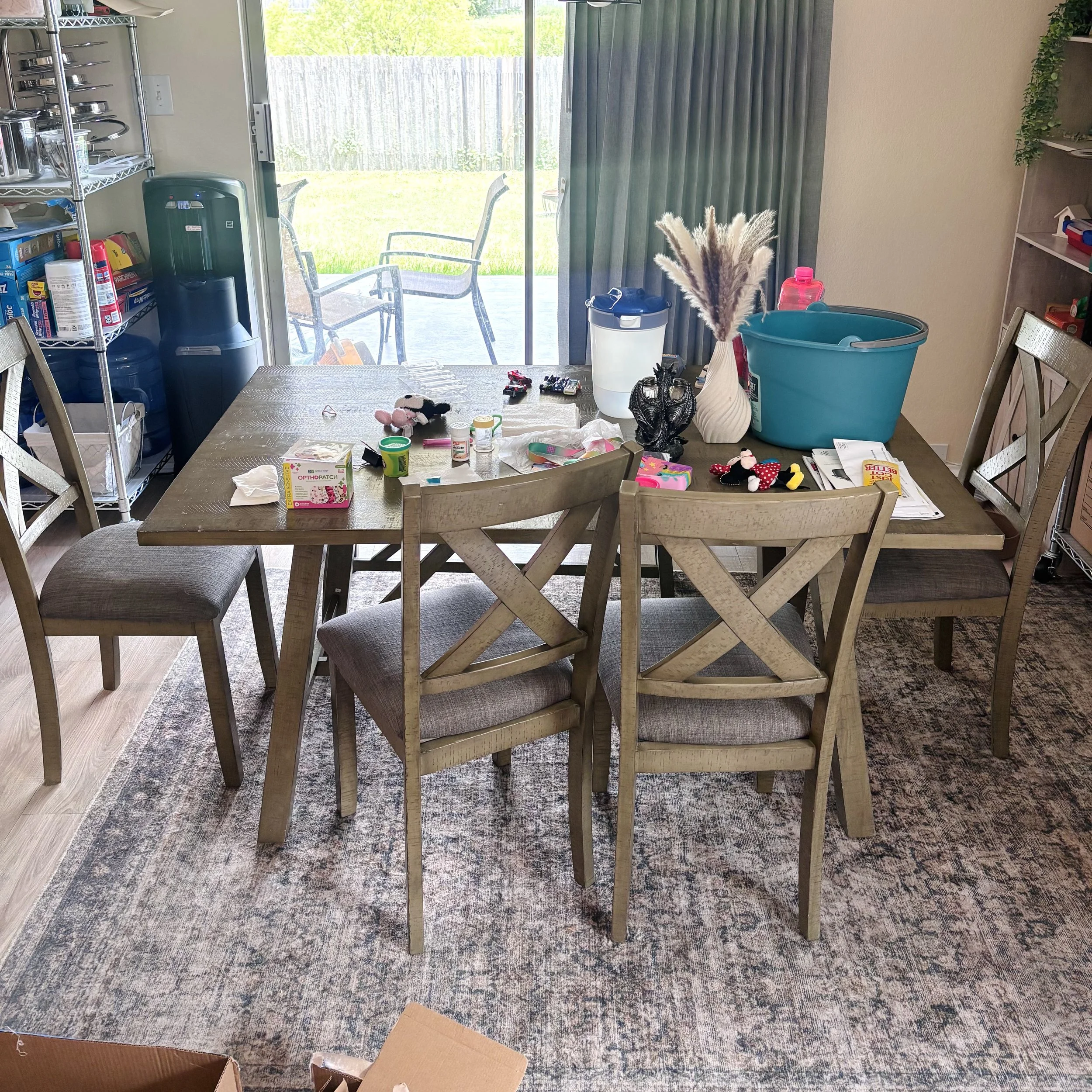 Inside view of a dining area with a table and chairs, cluttered with various items including toys, containers, and papers, with a sliding glass door showing an outdoor patio with chairs and green grass.