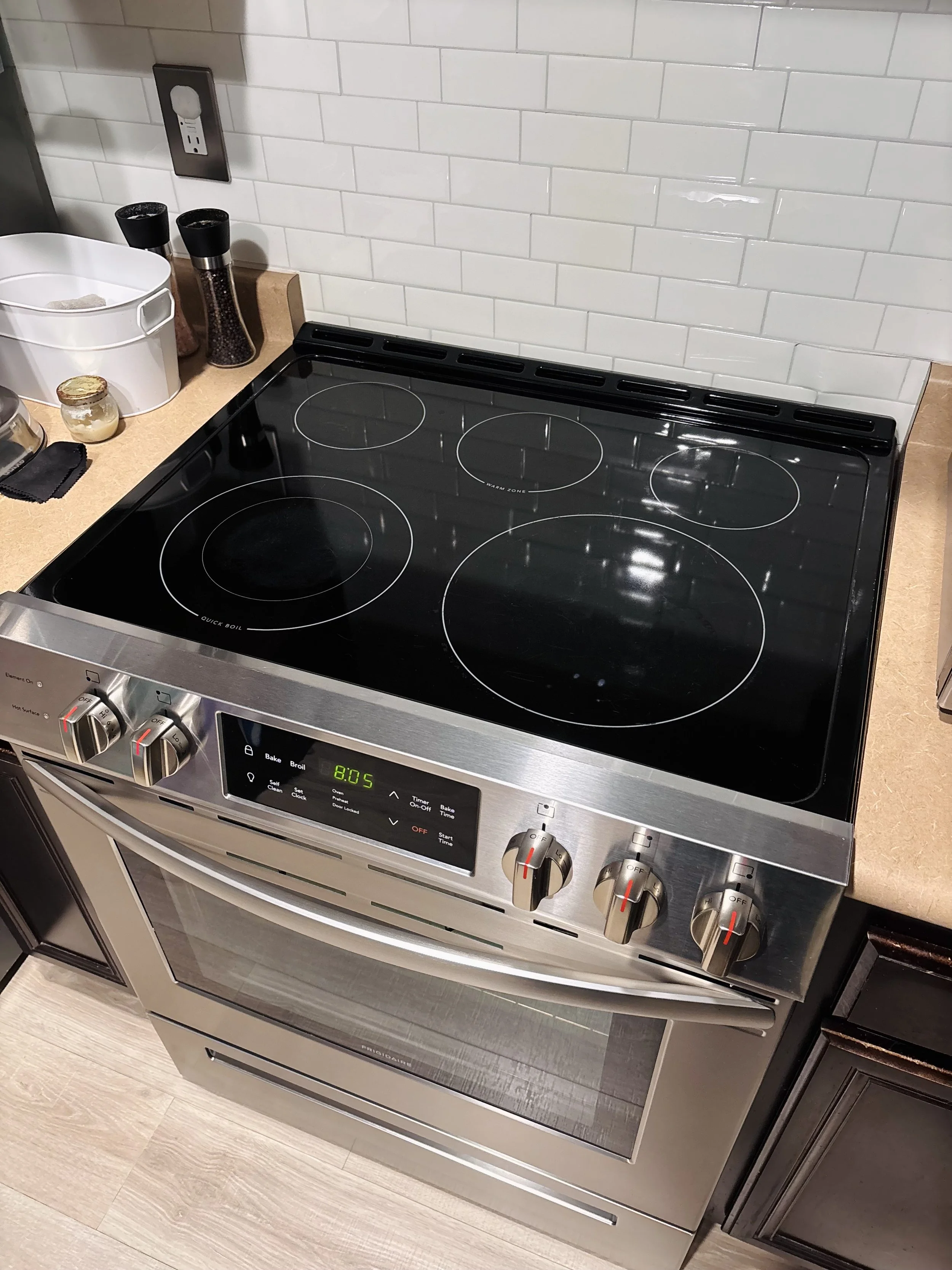 A modern stainless steel electric stove with four coil burners and control knobs. The stove has a digital clock display showing 2:05. It is in a kitchen with a light-colored wooden floor and a white tiled backsplash.