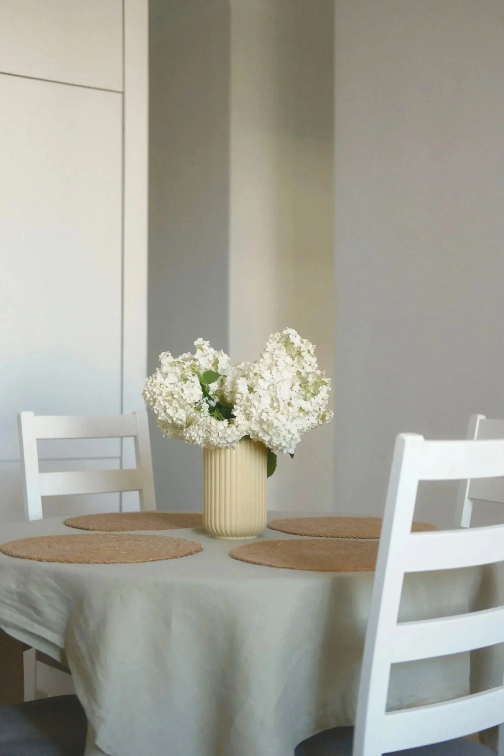 A white table with a beige tablecloth, set with a vase of white hydrangeas and four round woven placemats. Four white chairs surround the table, and the background features plain light-colored walls.