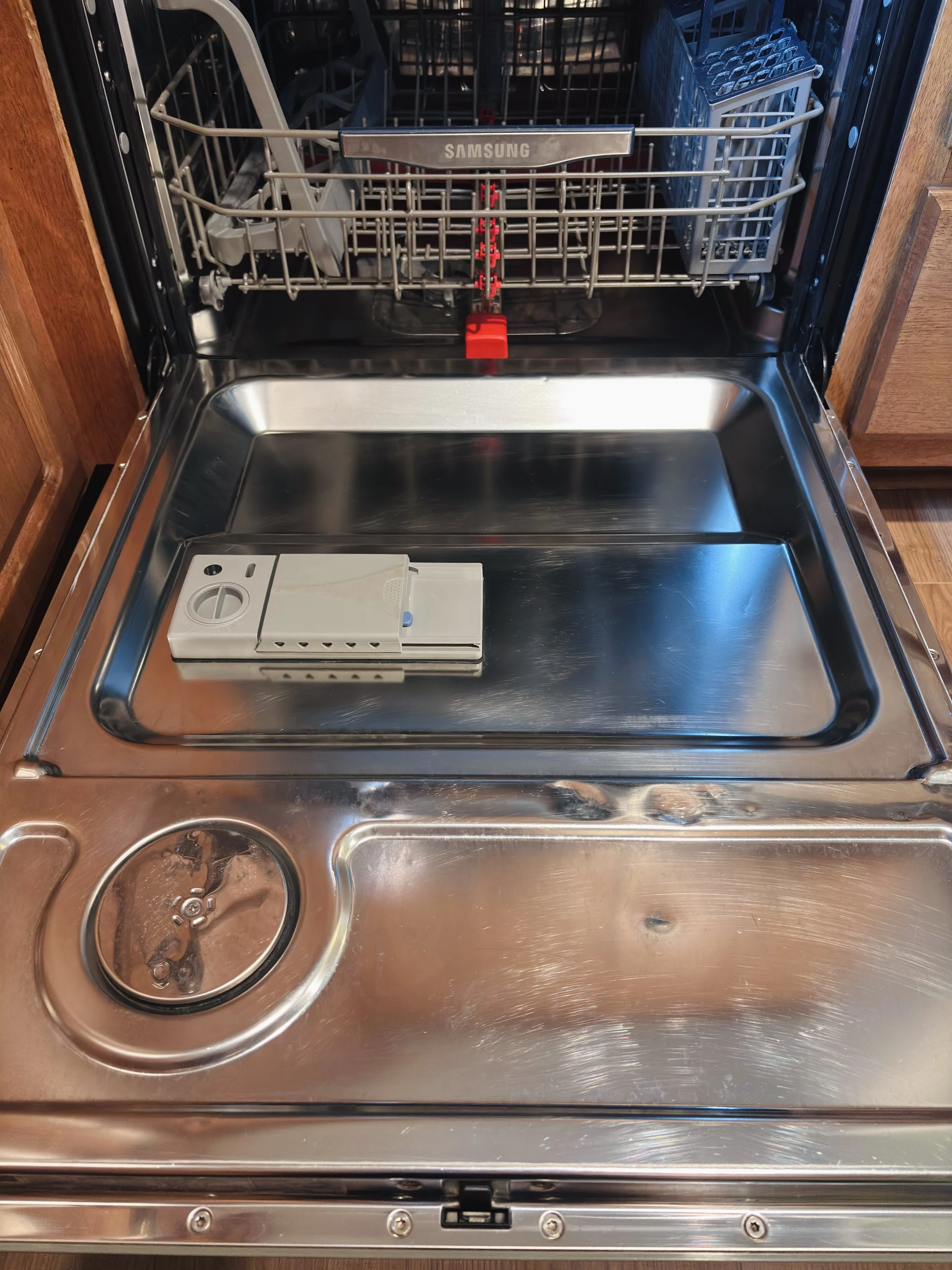 An empty dishwasher with stainless steel interior, a small white container at the bottom right, and a rack with a gray plate holder, with a wood cabinet to the sides.