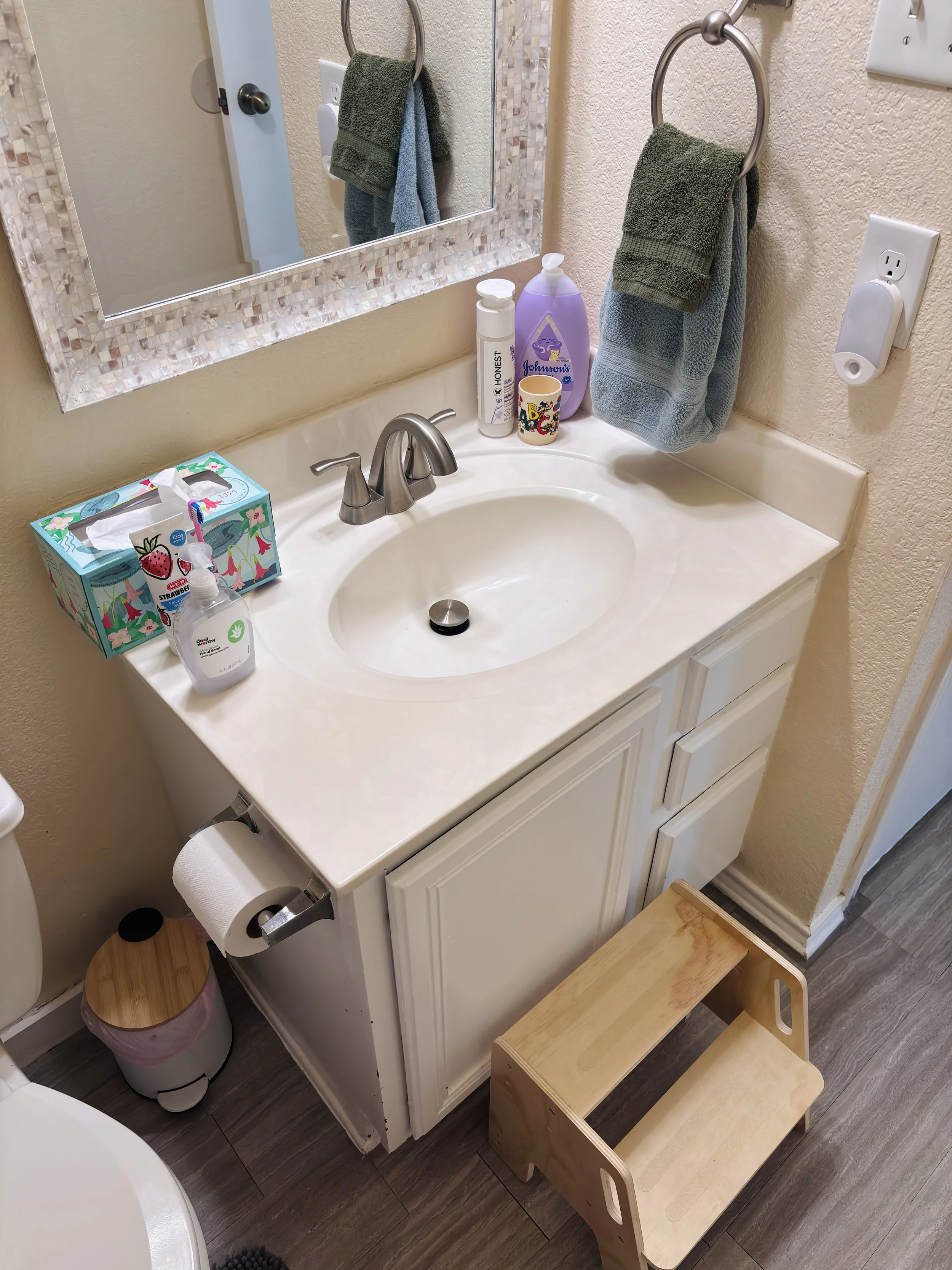 Bathroom vanity with a sink, mirror, hand soap, tissues, lotion, and towels hanging on ring hooks. A step stool is beside the vanity, and a trash can is visible underneath.
