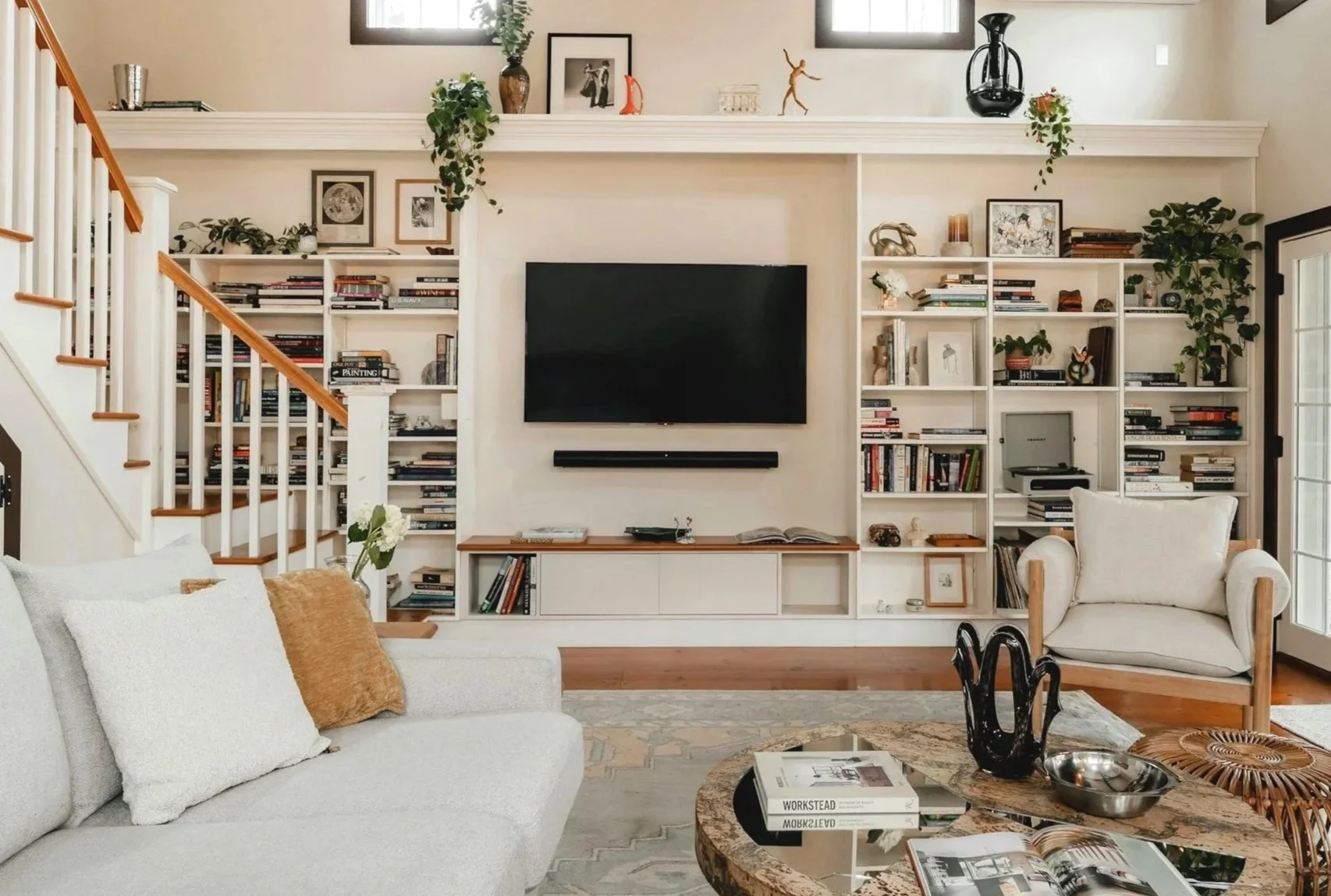 Living room with white sofa, beige cushions, and a coffee table with magazines and decorative items. Built-in white bookshelf with books and decor, black flat-screen TV mounted on the wall, and an armchair with a pillow. Large windows on either side of the TV wall and a staircase with wooden handrails on the left.