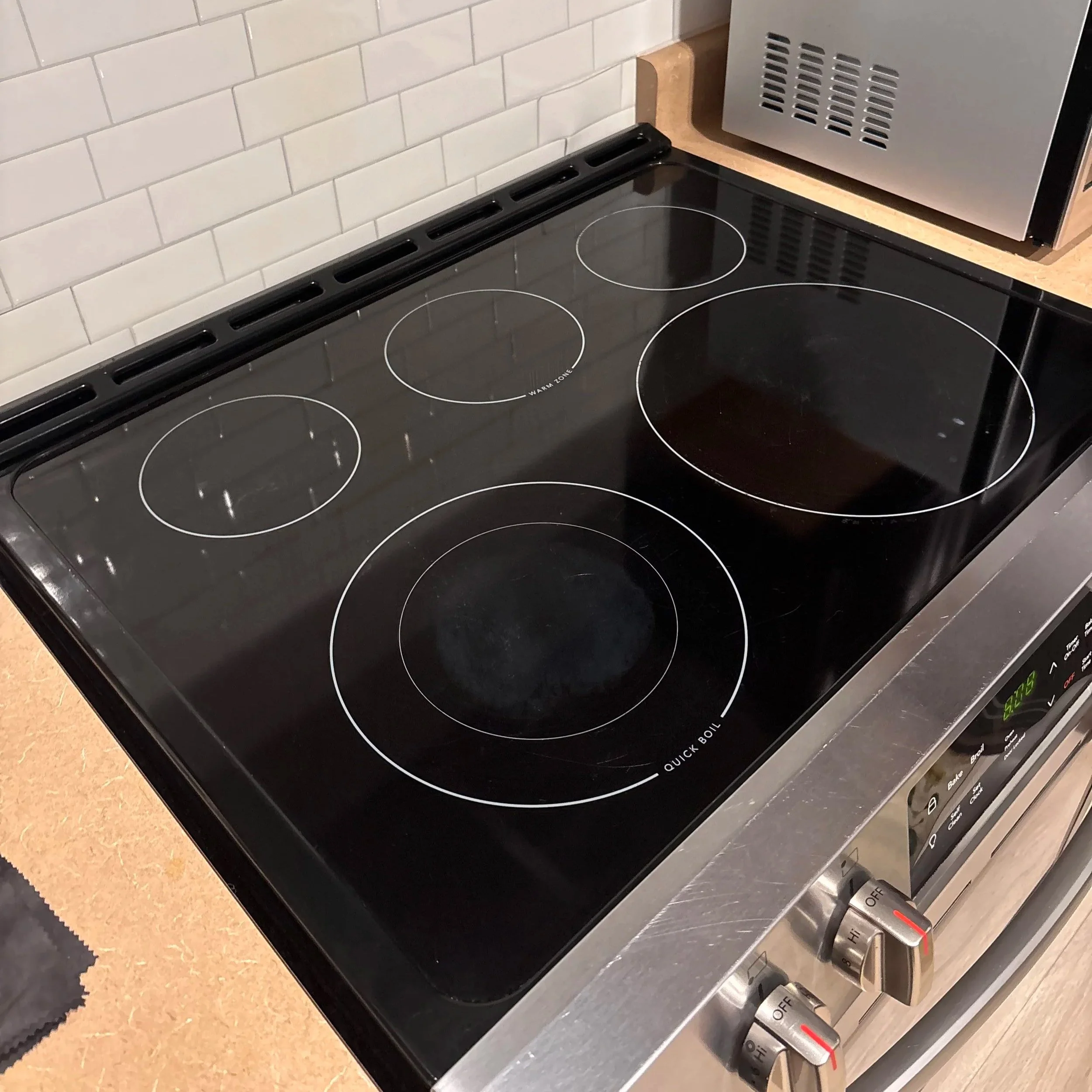 A black electric stovetop with white outlined circular burners, a control knob on the front panel, and a white-text label that reads 'WARM ZONE,' situated against a tiled kitchen wall with a stainless steel appliance nearby.
