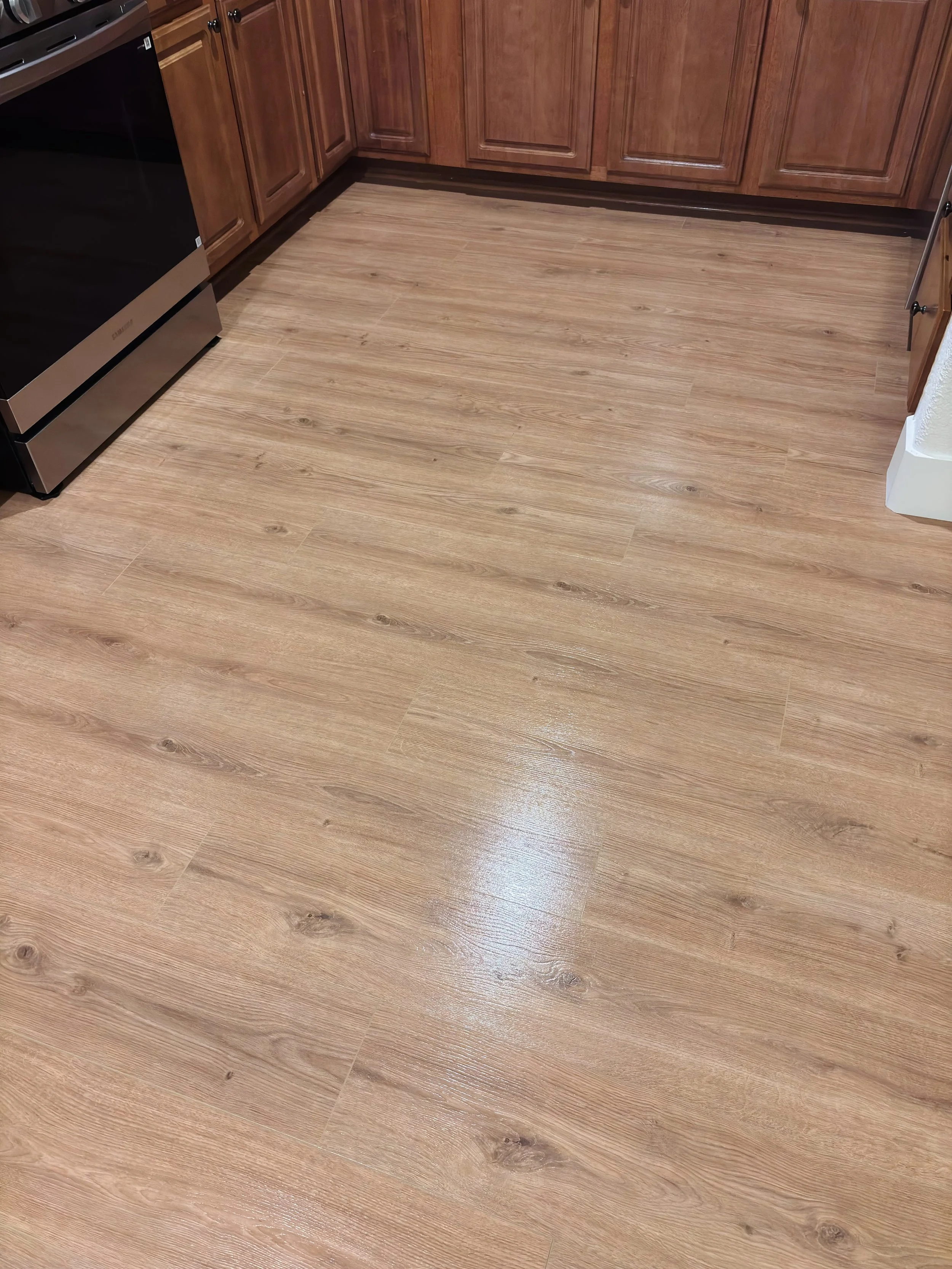 A kitchen with wooden cabinets and a black and stainless steel oven or stove. The floor is a light wood laminate. Part of the wall and a corner of the room are visible in the image.