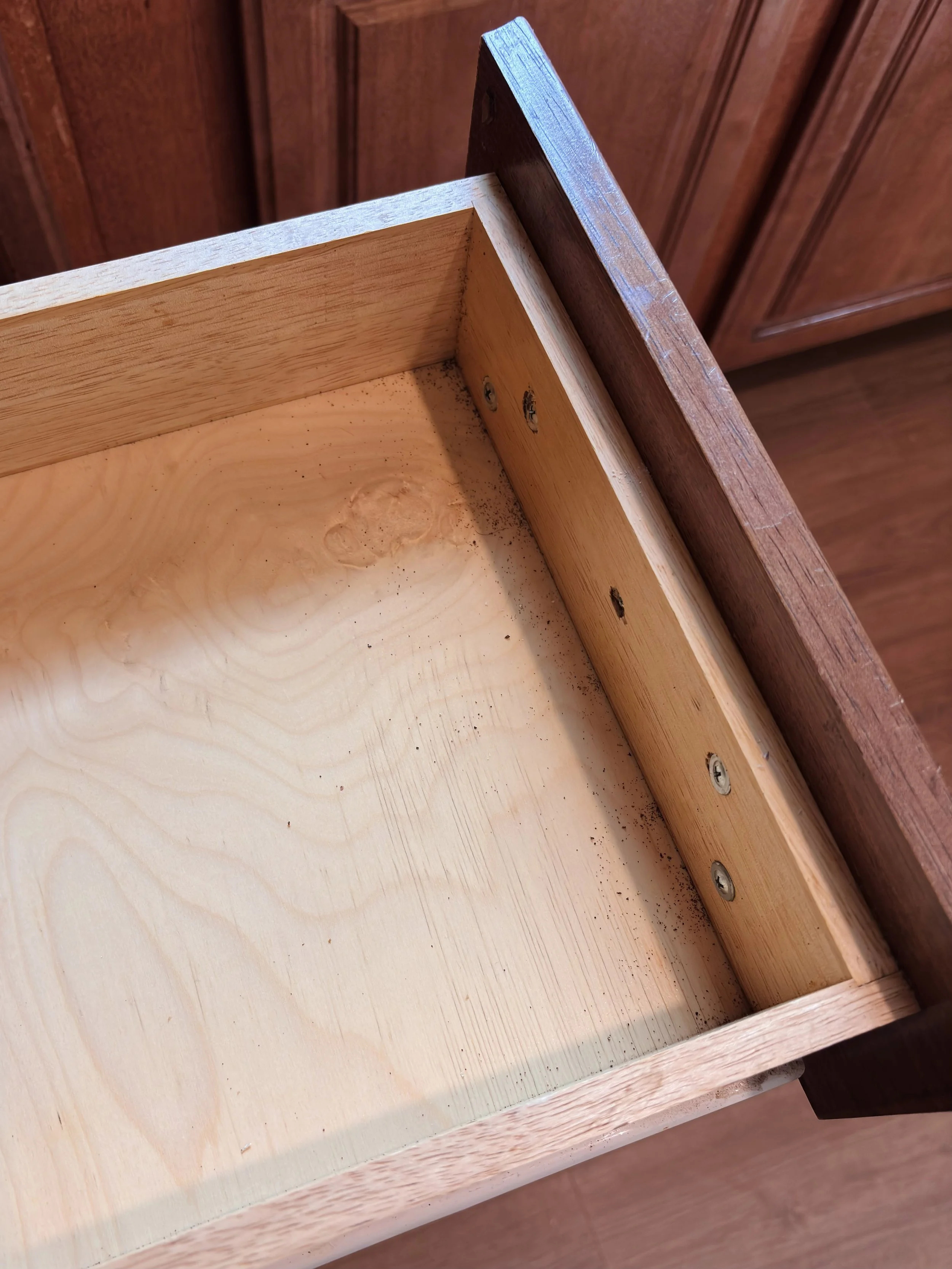 Close-up of the inside corner of a wooden drawer with metal screws, showing the layers of wood and some dust or small debris.