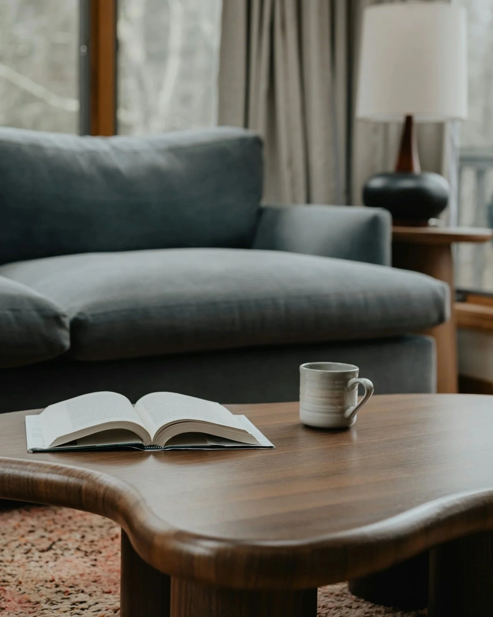 Open book and ceramic mug on a wooden coffee table in a cozy living room.