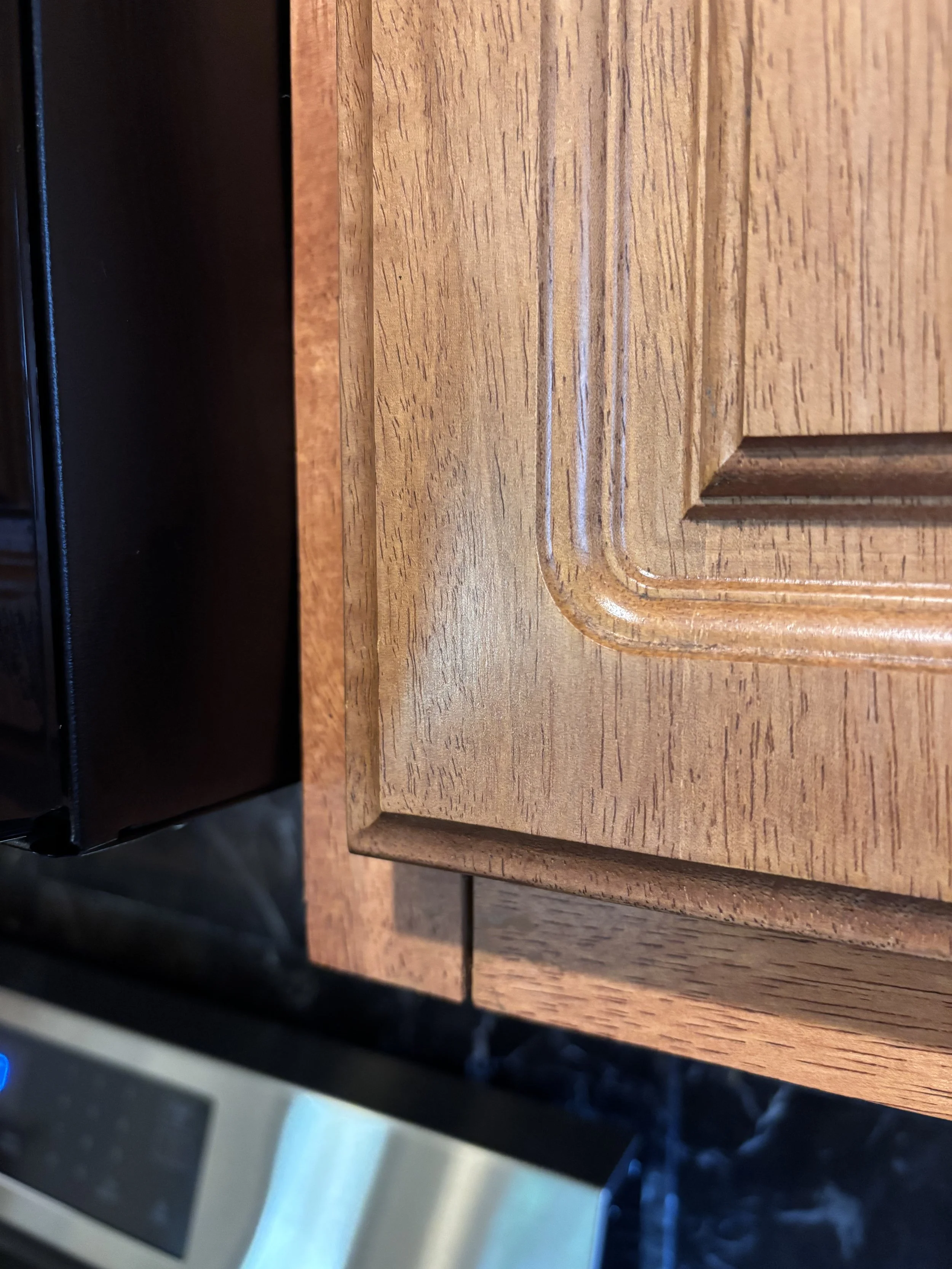 Close-up photo of a wooden cabinet with detailed grain and a rounded edge, with a black object and part of a microwave oven in the background.