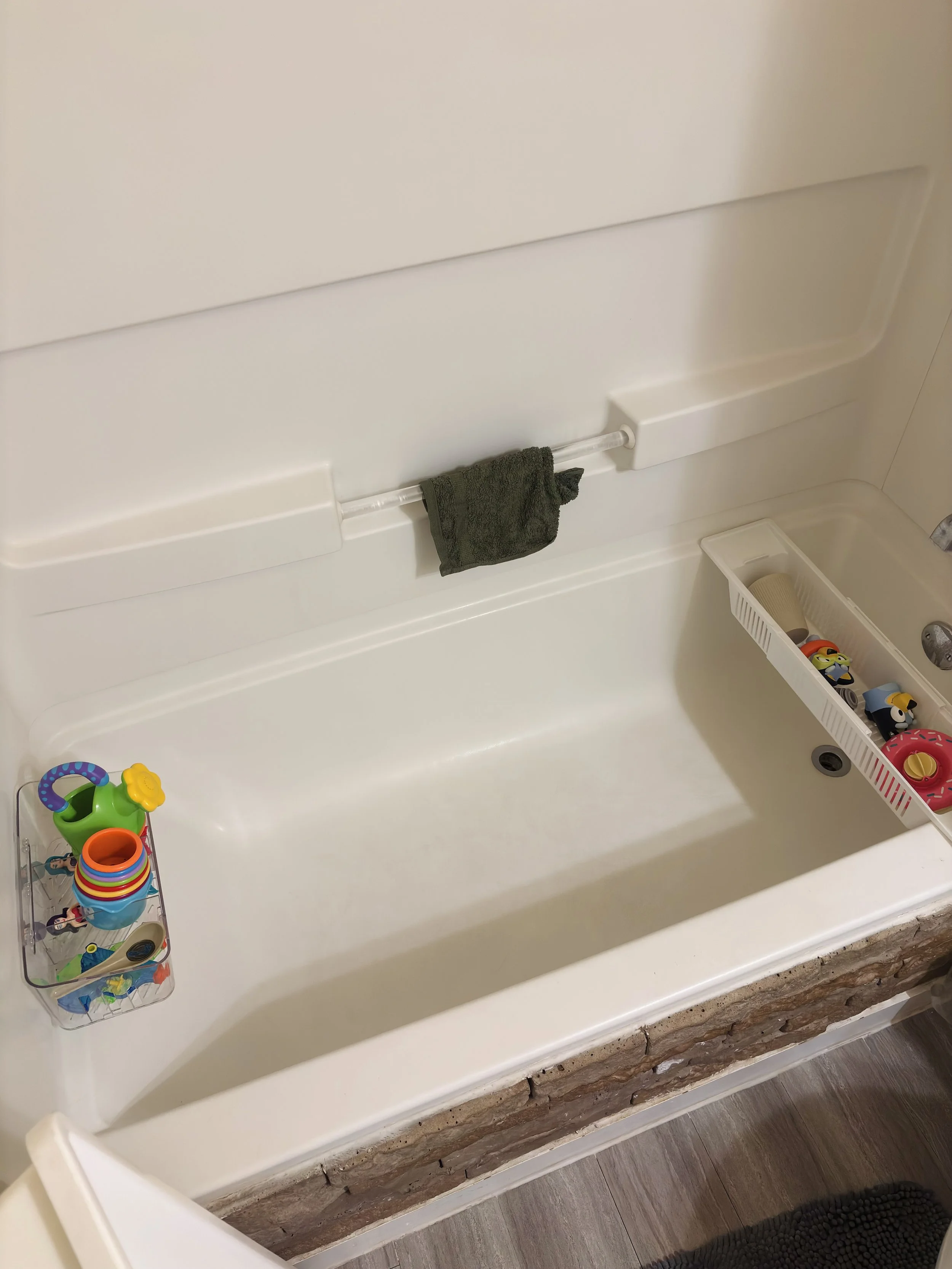 Bathroom bathtub with a small green towel hanging from a bathtub rail, a white plastic basket on the edge containing toy cars and a brush, a small clear container with colorful toys, and part of a rug on the wooden floor.