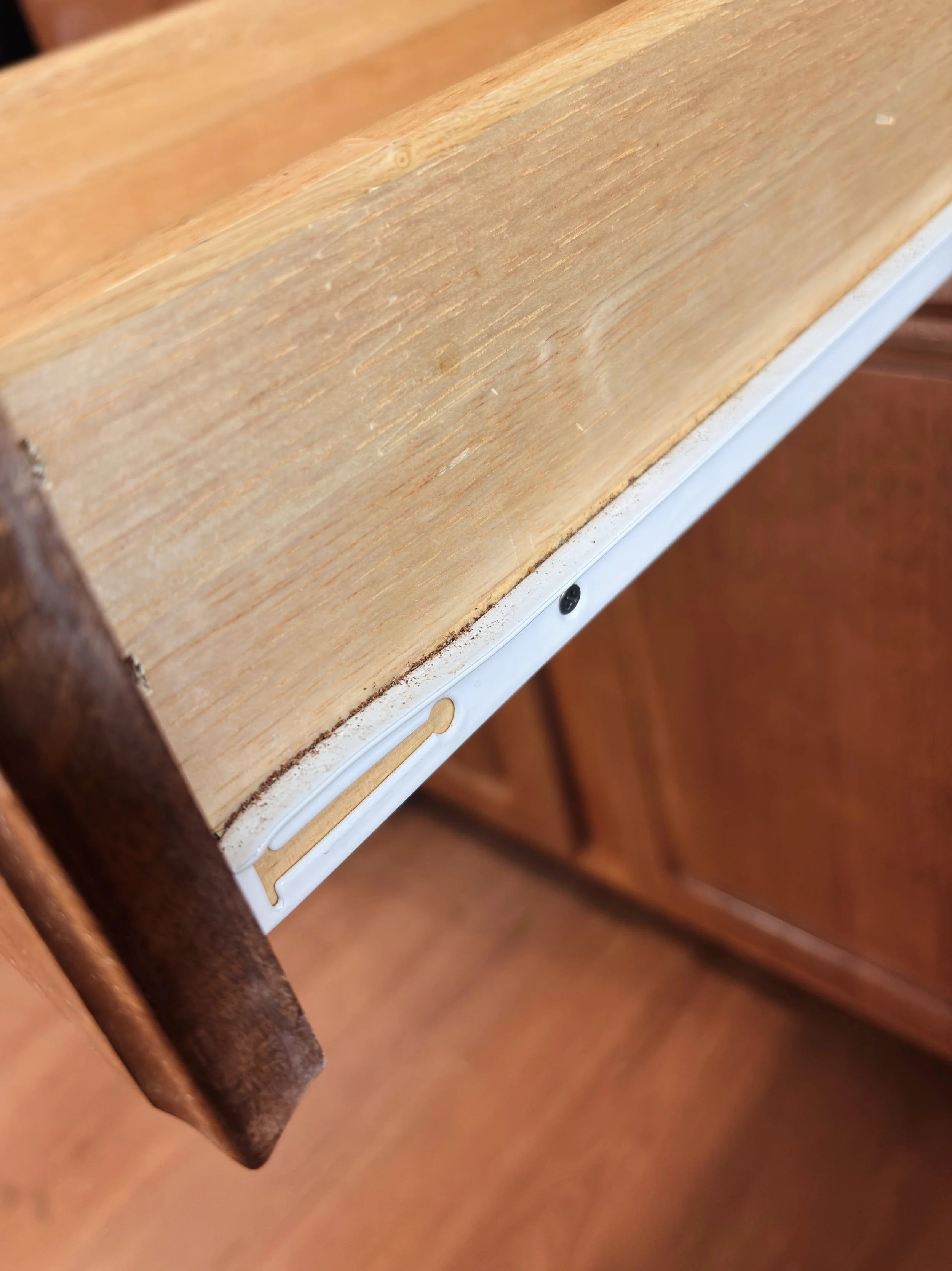 Close-up of the edge of a wooden piece on a piece of furniture, possibly a drawer or a bed frame, with a white metal support underneath and a screw visible.