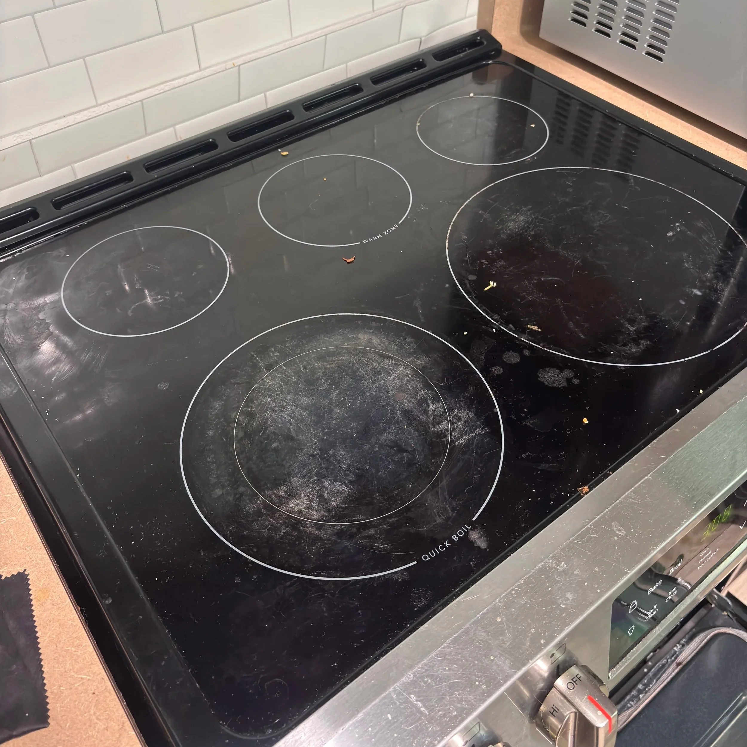 A dirty black glass electric stove with four burners, surrounded by a beige countertop. There is a control panel with buttons and knobs at the front, and a gray microwave oven to the right. The kitchen has a white tiled wall behind the stove.