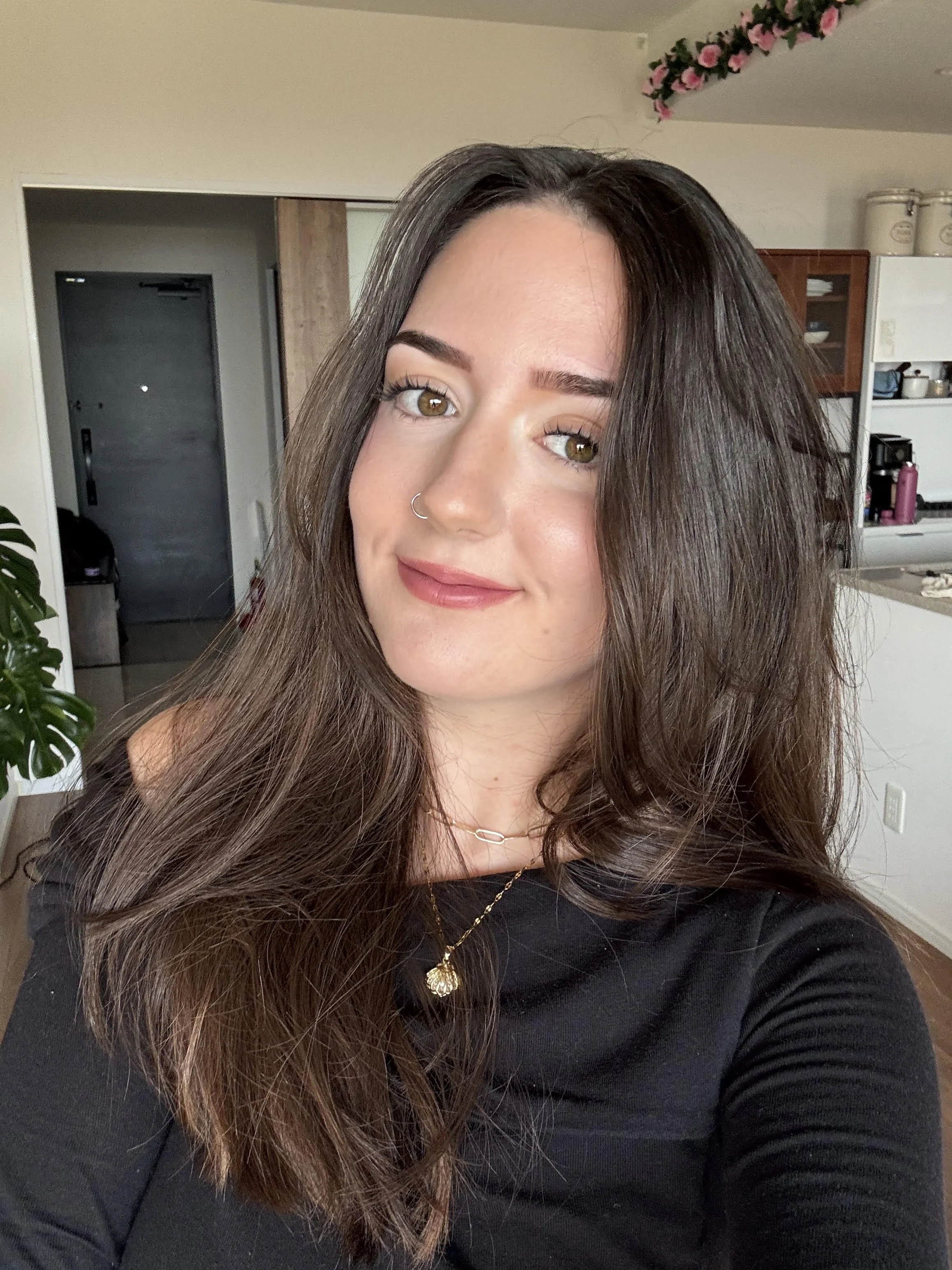A woman with long brown hair, hazel eyes, and a nose ring taking a selfie indoors. She is wearing black clothing and jewelry, including a gold necklace with a heart-shaped pendant. In the background, there's a kitchen with shelves, a door, and decorative pink flowers on the ceiling.