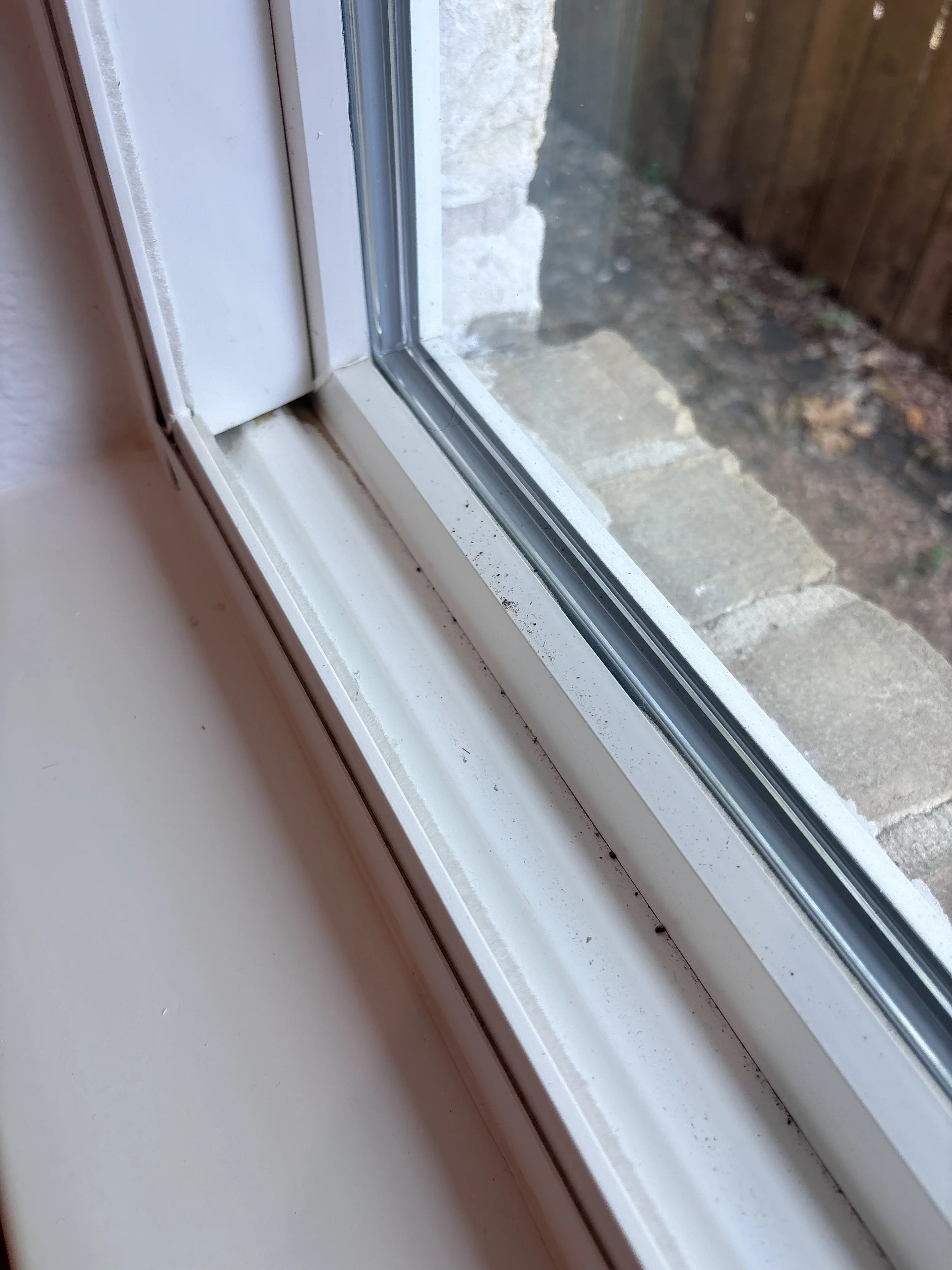 Close-up of a window sill with dust and dirt, showing a portion of a glass window and a wooden fence outside.