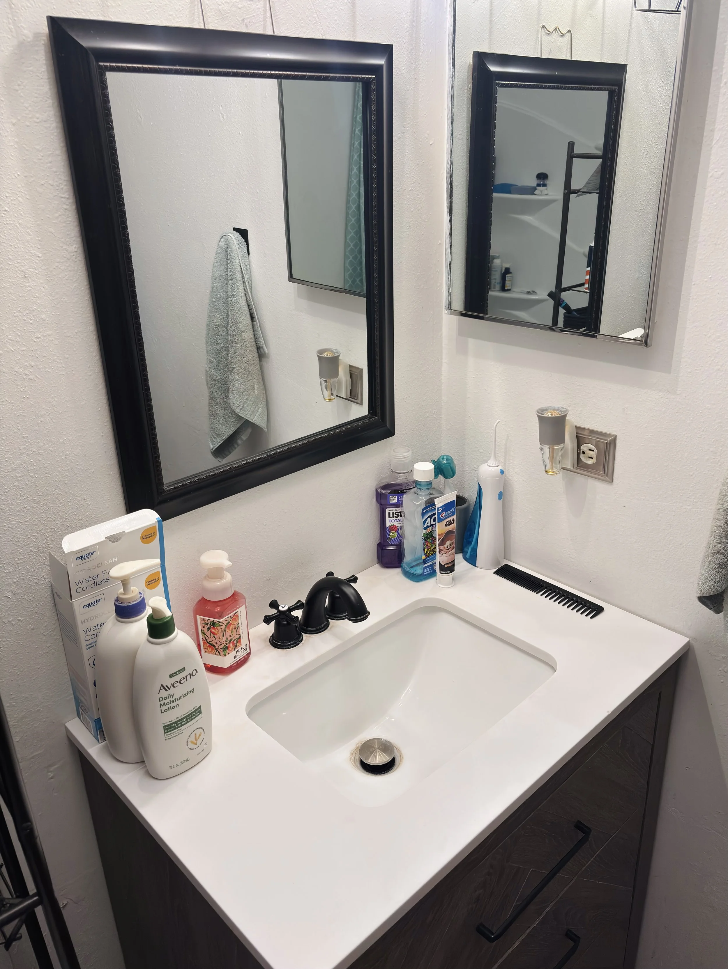 Bathroom vanity with various toiletries, two mirrors, a towel hanging on a hook, and a black faucet in the sink.