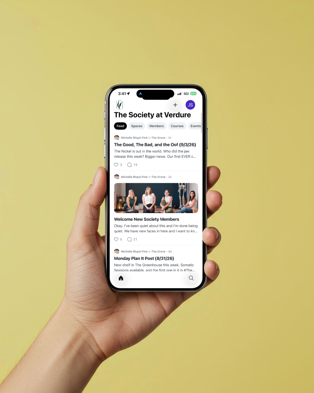 A person holding a smartphone showing a social media group page called 'The Society at Verdure' with posts about meetings and new members, against a yellow background.