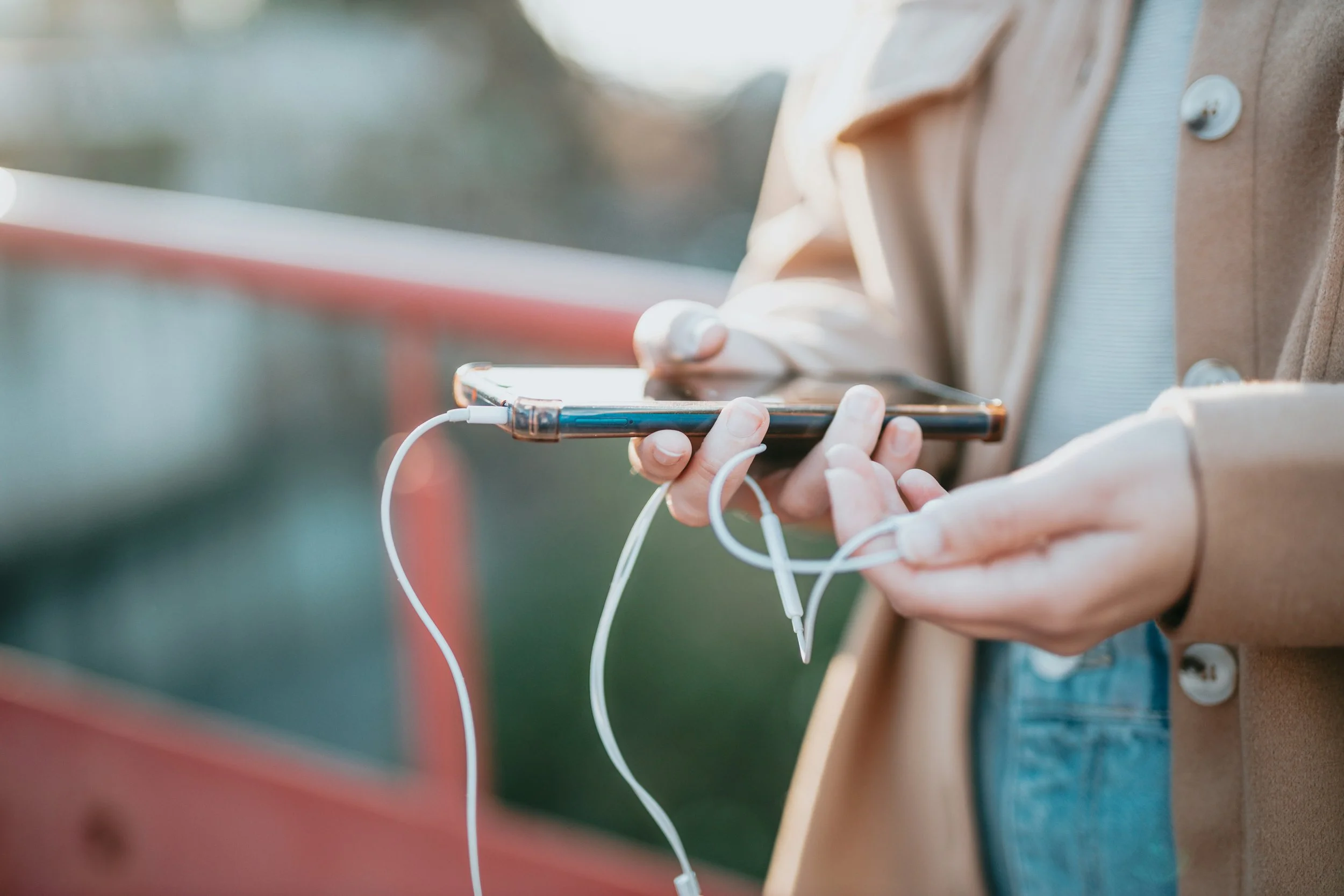 Close-up of a person holding a smartphone with earbuds connected, standing outdoors near a colorful wall.