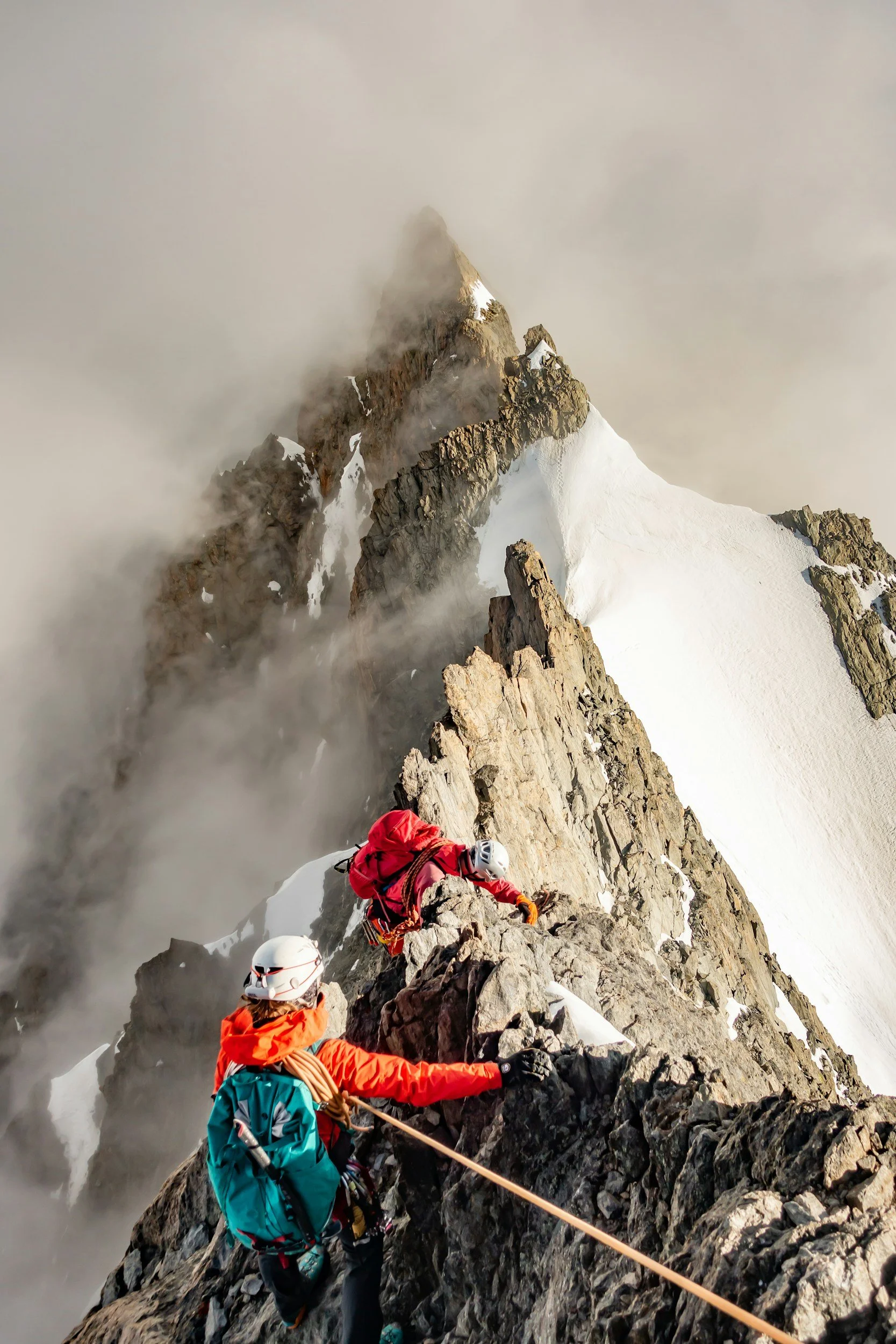 Mountain climbers ascending a snow and rock ridge on a high peak, with clouds and fog surrounding the summit.