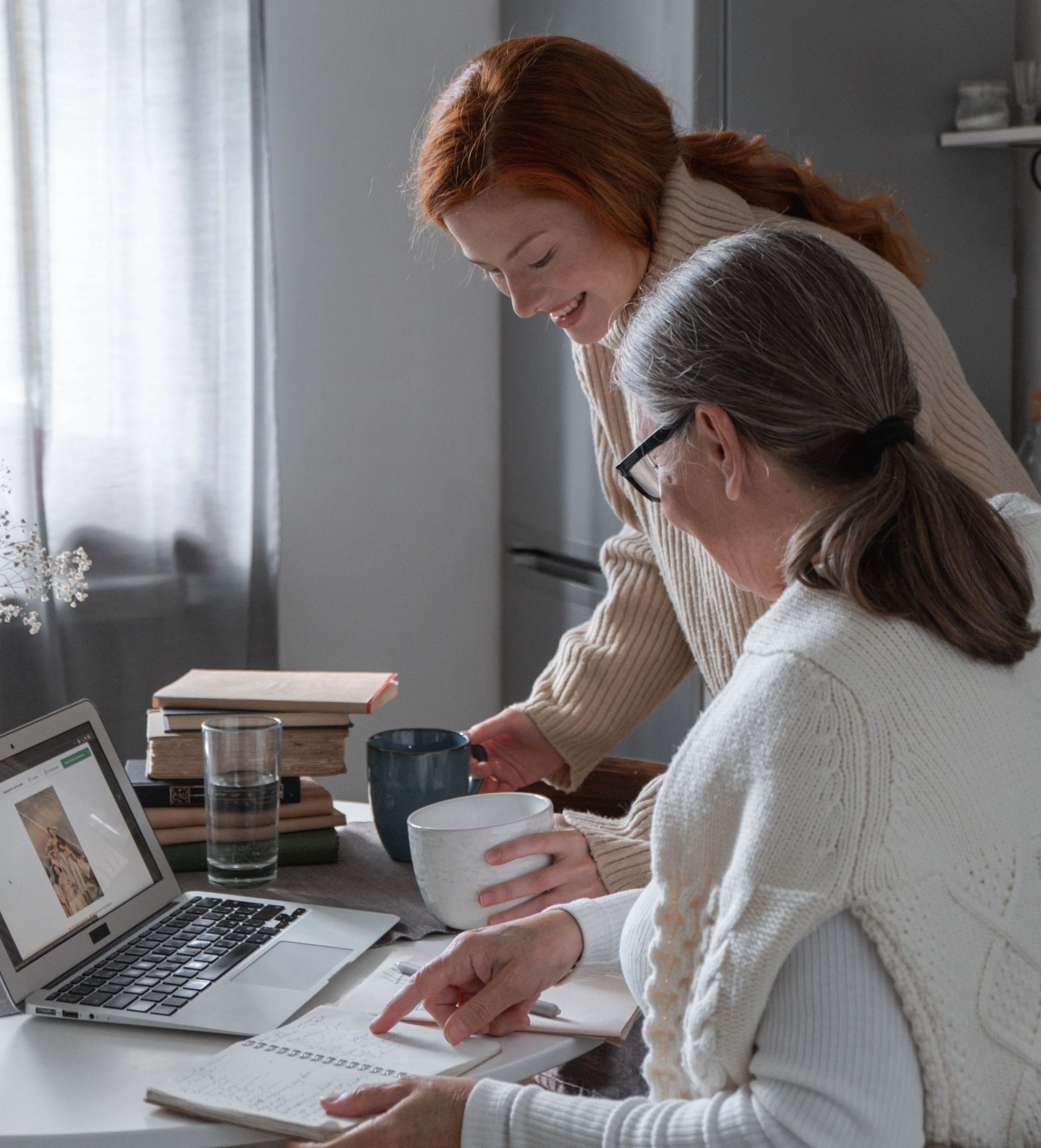 Two women are sitting at a table, engaged in a discussion. One woman with reddish hair is smiling and leaning forward, while the other with gray hair and glasses is pointing at notes in a notebook. The table has books, a laptop, gift boxes, a glass of water, and a mug. The room has natural light coming through curtains.