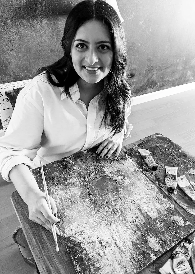 A smiling woman sitting at a table with art supplies, including paint tubes and brushes, in a room with a textured wall.