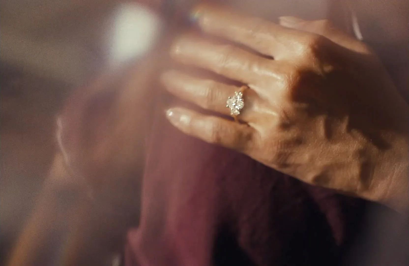 A close-up of a hand with a large, sparkling diamond ring on the ring finger, resting on a soft, pink fabric.