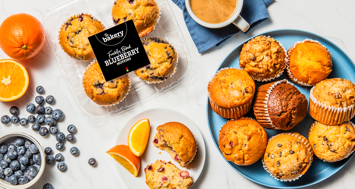 Assorted muffins on a blue plate, a container of blueberry muffins, fresh blueberries, orange slices, and a cup of coffee on a white surface.