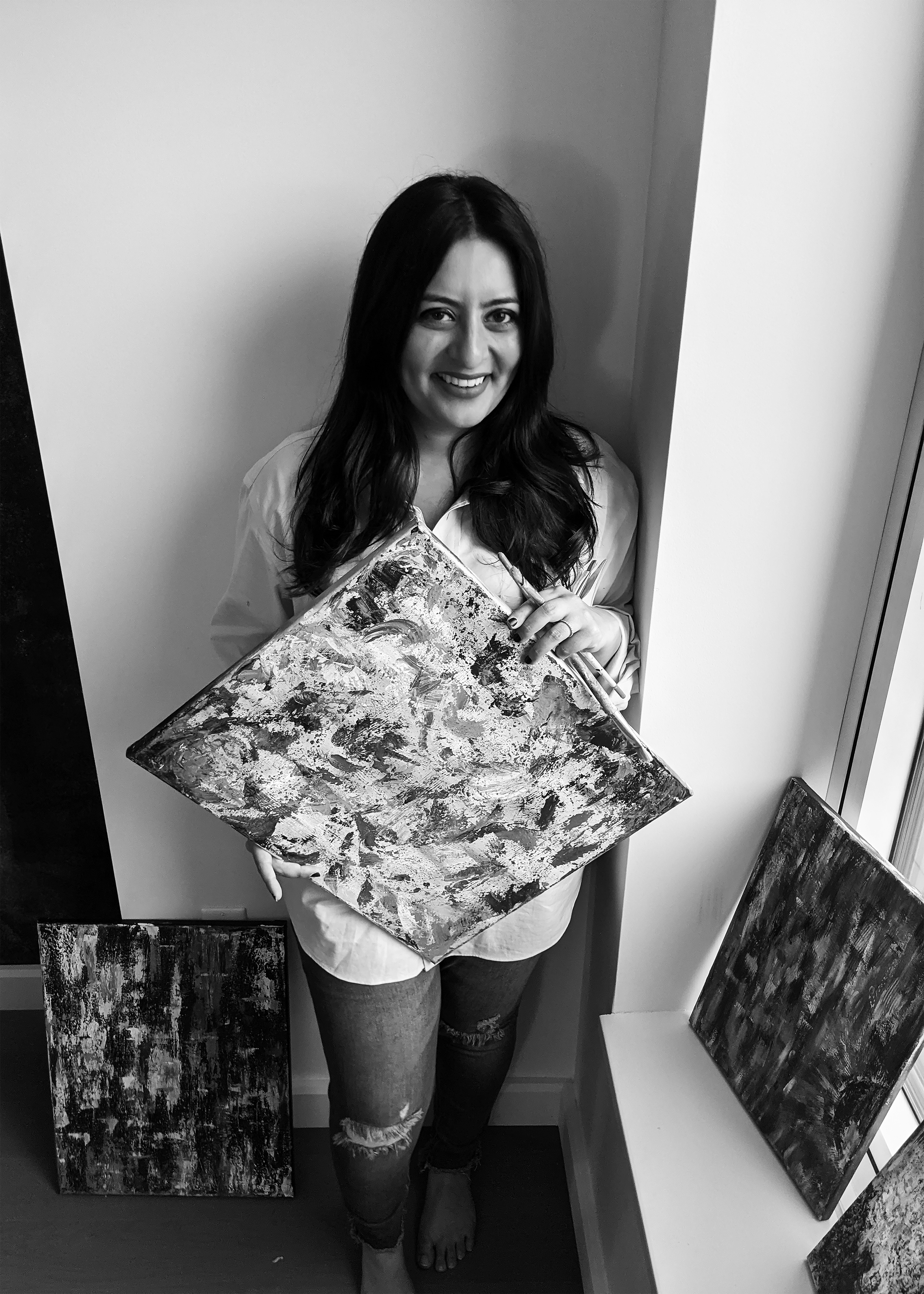 A woman with long dark hair smiling, holding a paintbrush and an abstract painting, standing in front of a wall with her lower legs visible, wearing ripped jeans and a light-colored shirt.