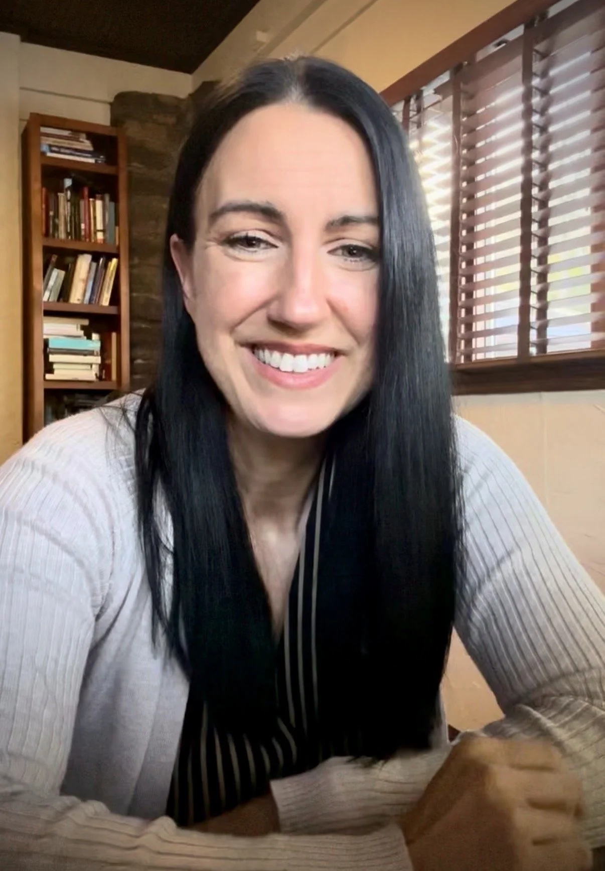 A woman with long dark hair smiling at the camera, sitting indoors near a window with wooden blinds, with a bookshelf filled with books in the background.