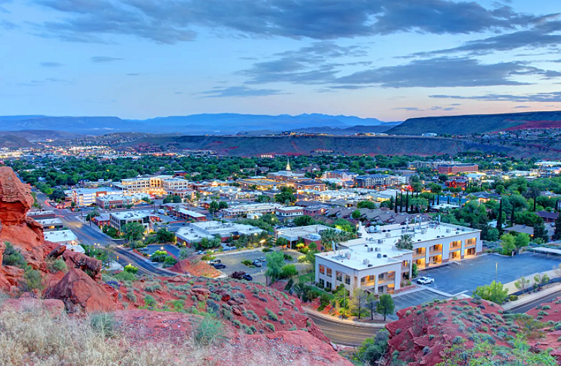 A panoramic view of a city in a valley with mountains in the background, taken from a hillside with red rocks and sparse desert vegetation.
