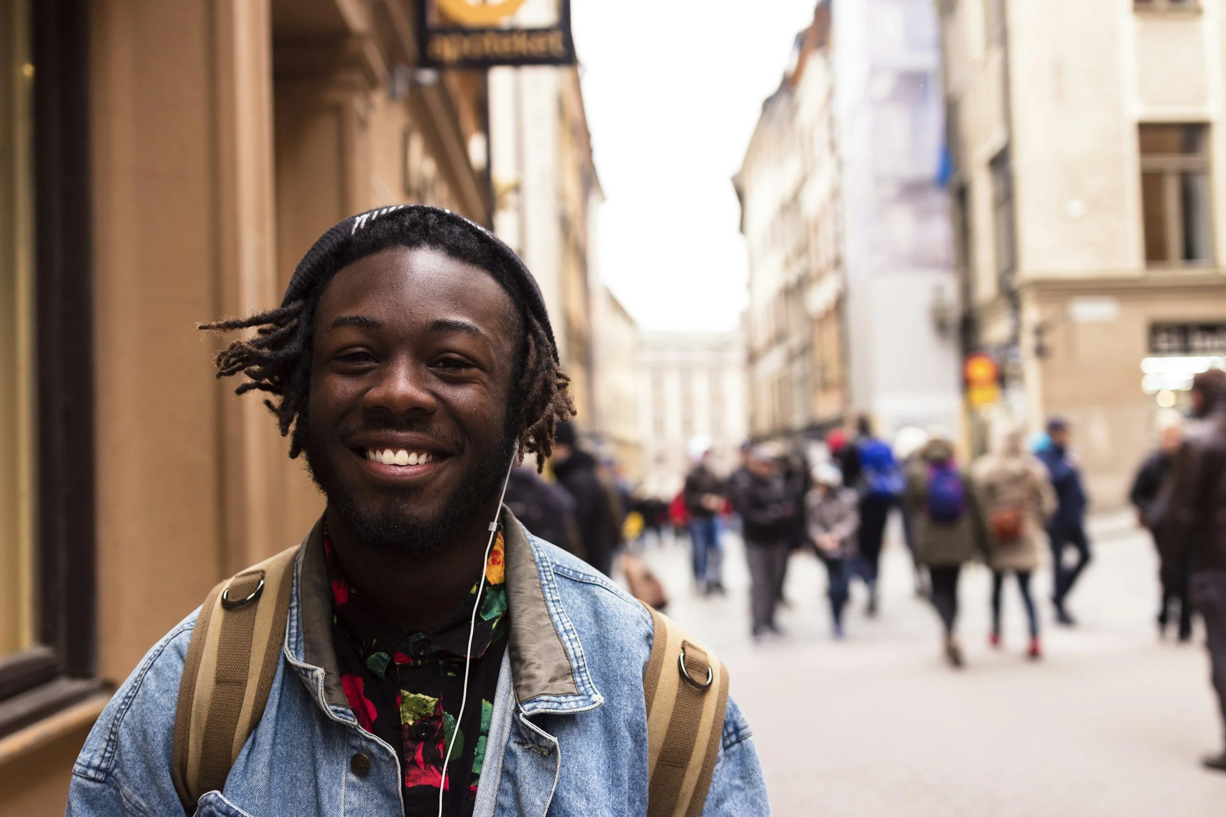 A young man with dreadlocks smiling, wearing a denim jacket, a colorful shirt, a beanie, and carrying a backpack, standing on a busy city street with buildings and people in the background.