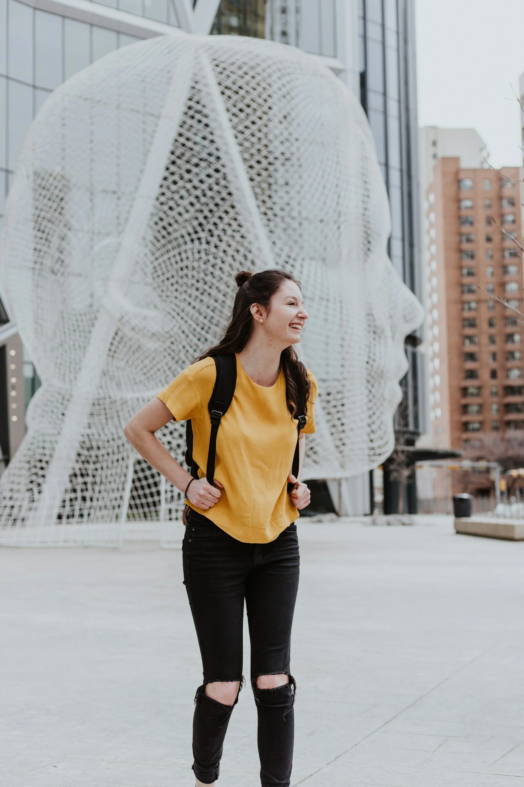 A young woman with long dark hair, wearing a yellow t-shirt, ripped black jeans, and a backpack, smiling and standing outdoors in front of a large modern architectural structure resembling a human head sculpture, with city buildings in the background.