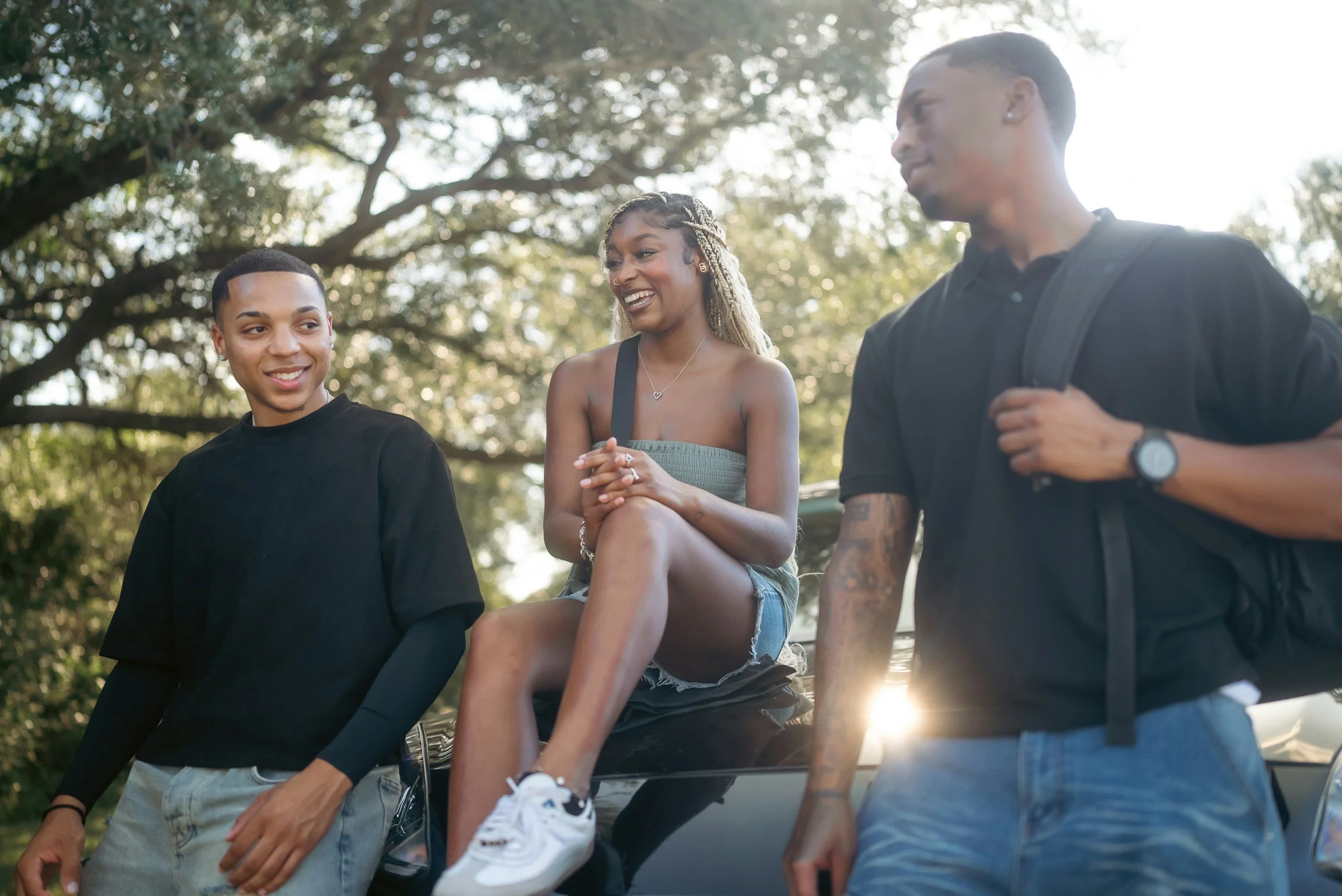 Three young adults, two men and one woman, are outdoors and appear to be enjoying a casual conversation. The woman, sitting on a car, is smiling and wearing denim shorts, a tank top, and sneakers. One man, standing to her right, has tattoos on his arm and is wearing a black T-shirt and watch. The other man, to her left, is also smiling and wearing a long-sleeve black shirt and light-colored pants. Trees with sunlight filtering through the leaves are in the background.