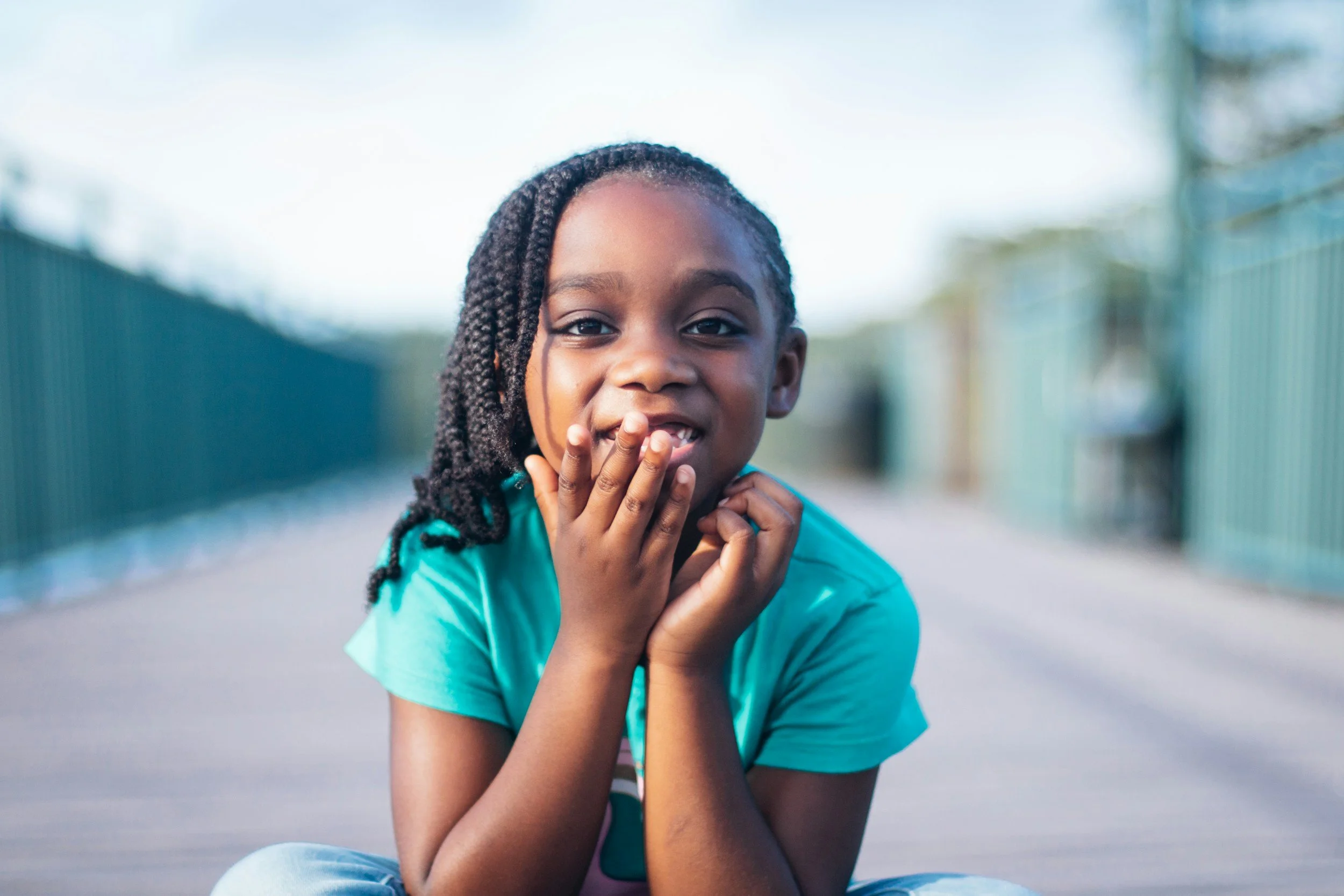 A young girl with braided hair smiling and touching her face while sitting outdoors on a pathway with a blurred fence and trees in the background.