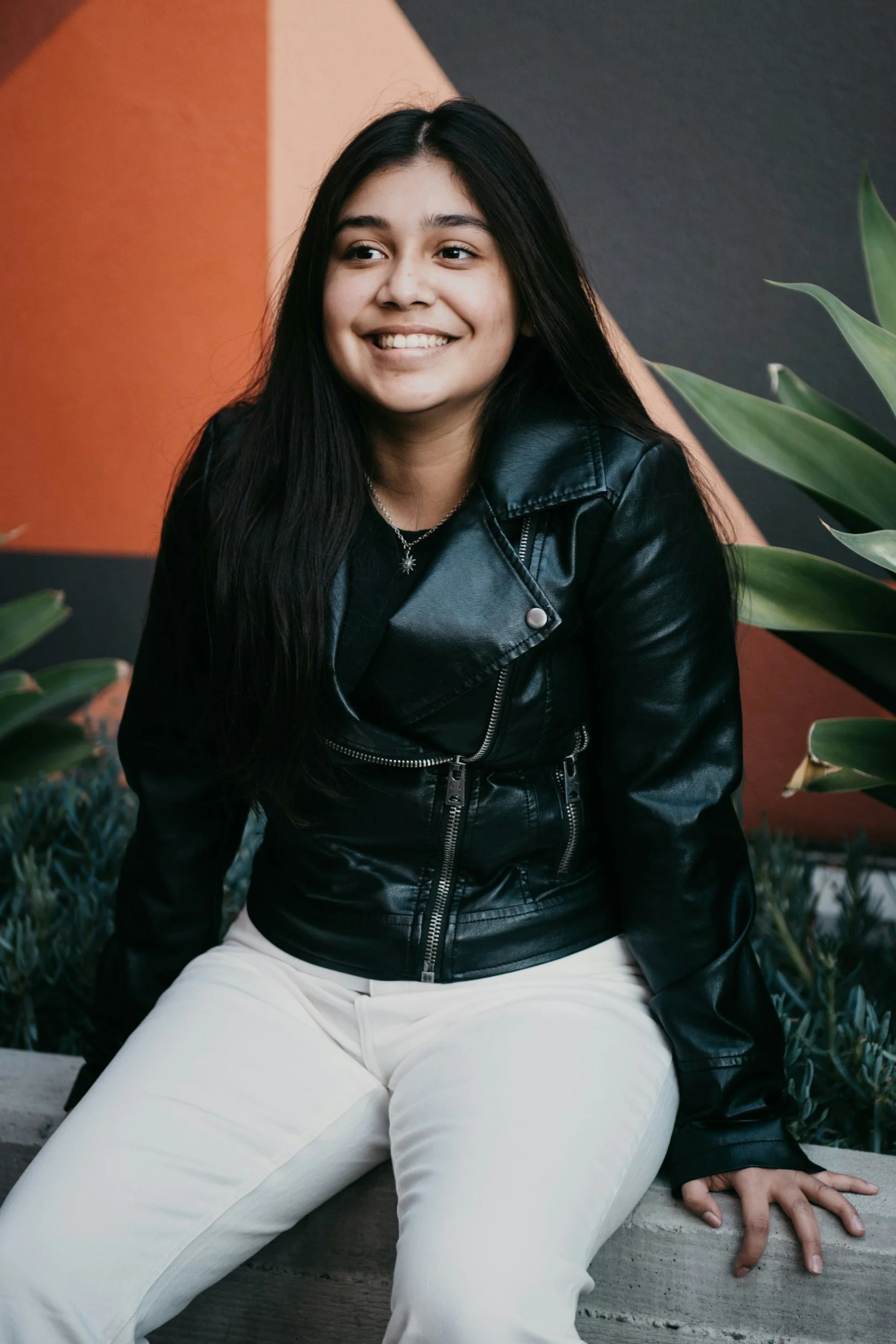 A young woman with long black hair, wearing a black leather jacket and white pants, sitting on a concrete ledge next to green plants, smiling and looking to her left.