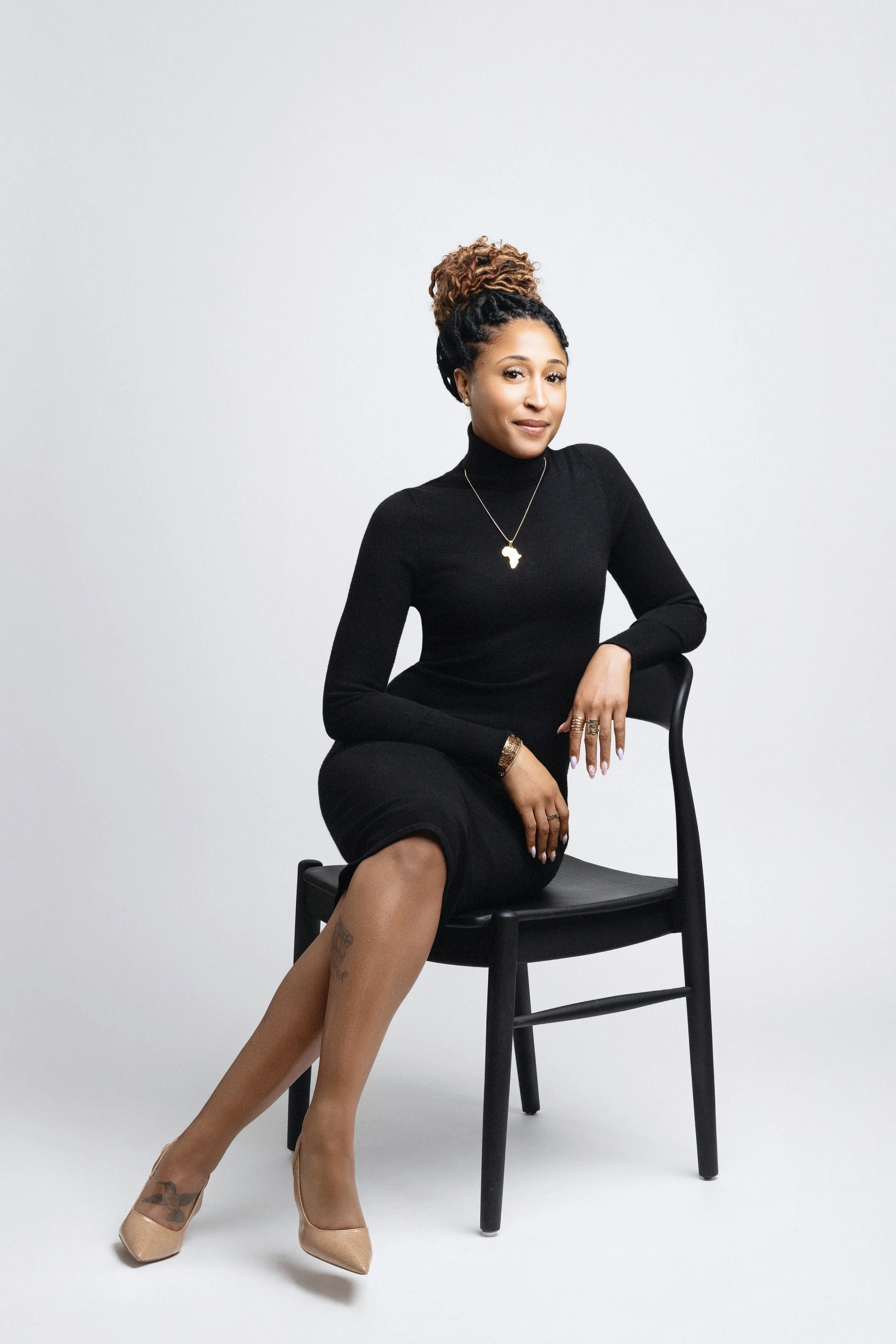 A woman with dark skin and curly hair styled in a bun with blonde highlights, wearing a black turtleneck and long black skirt, sitting on a black chair against a plain white background. She is accessorized with gold jewelry including a necklace with an Africa-shaped pendant, rings, and bangles, and is wearing nude high heels.
