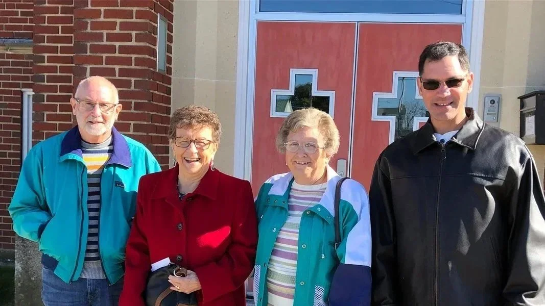 Four people standing outdoors in front of a building with a red door with a cross design. Two elderly women and two elderly men, smiling and dressed casually.