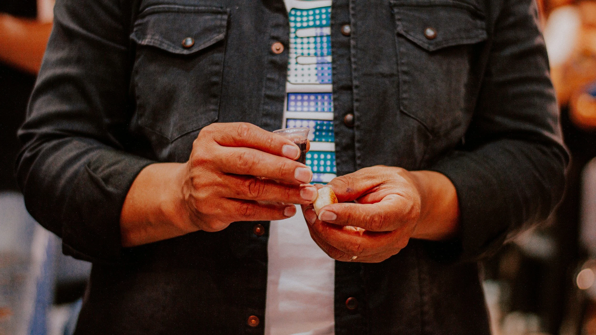 A person wearing a black jacket holding a small shot glass filled with dark liquid in one hand and a cookie in the other, in a social setting.
