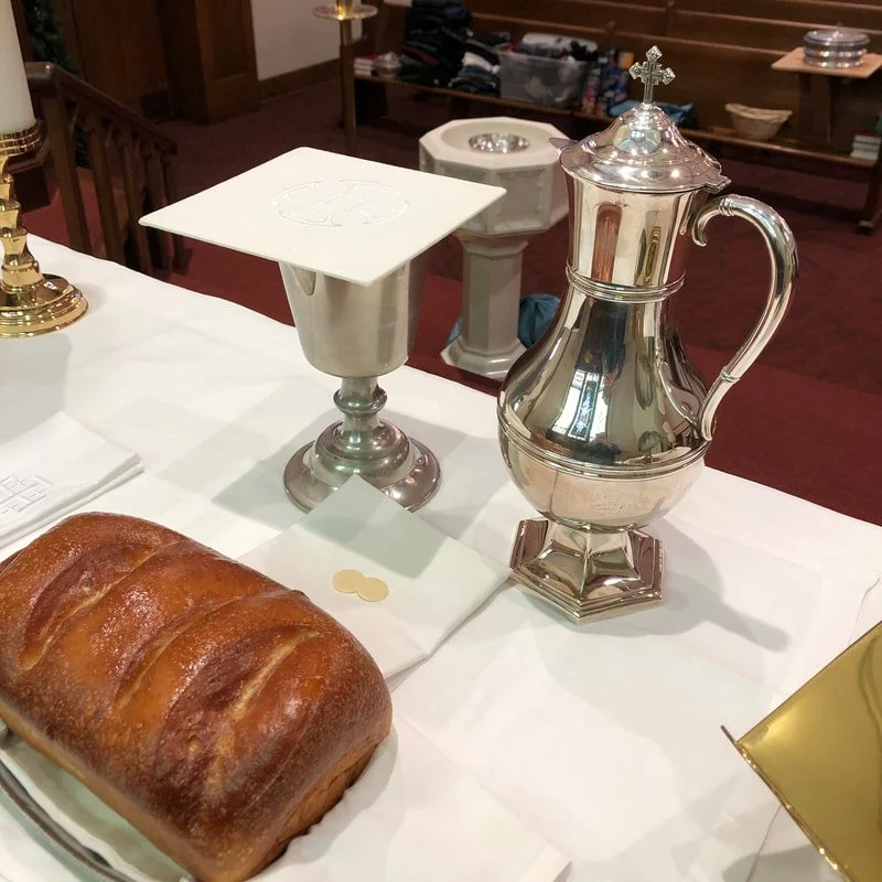 A loaf of bread, a silver candlestick holder, a silver vessel with a lid and cross on top, a small white cloth, and a napkin with a gold wax seal on a white tablecloth in a church setting.