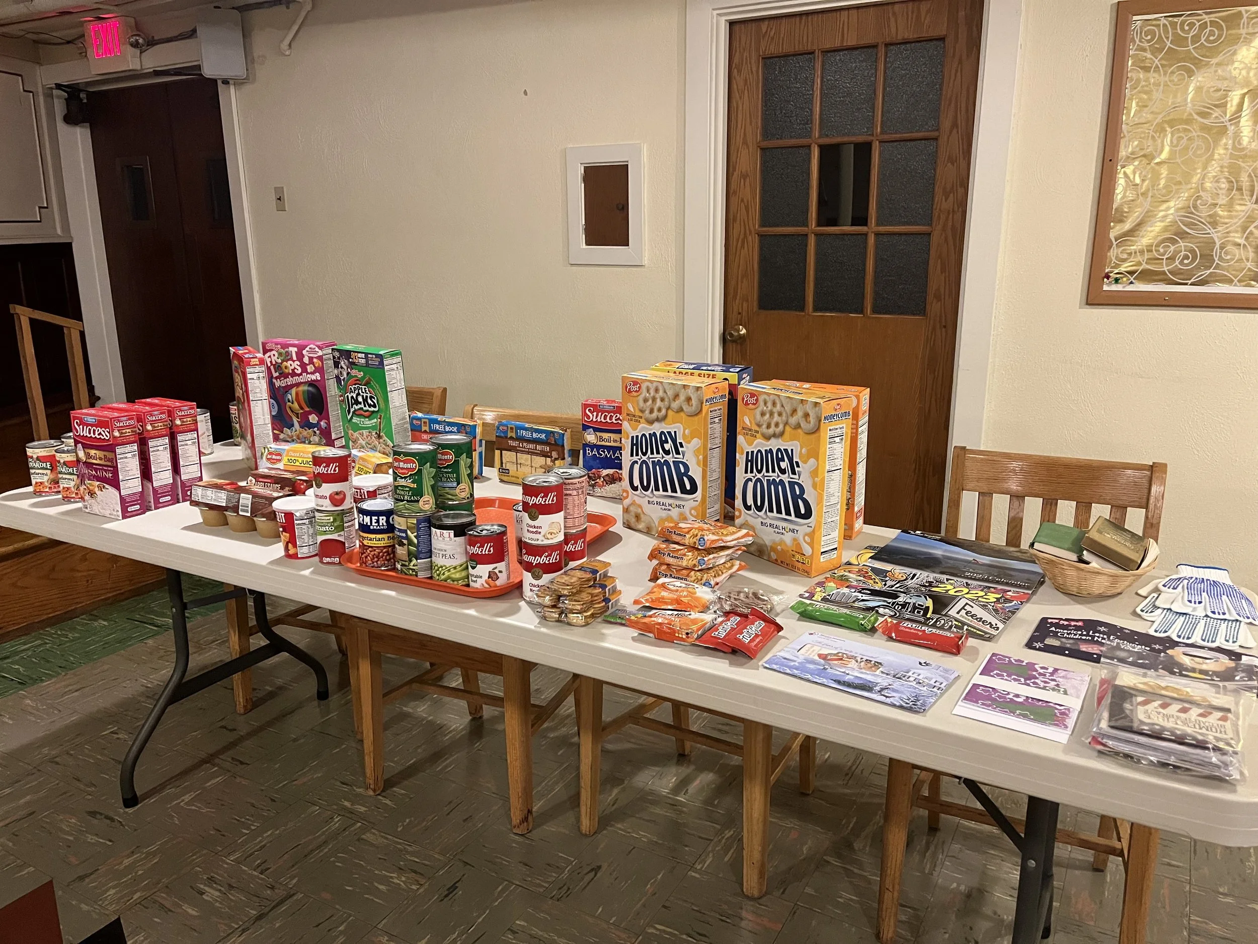 A long table covered with various food items, canned goods, cereals, and magazines inside a room with beige walls, a wooden door, and a bulletin board. The breakfast Blessing table