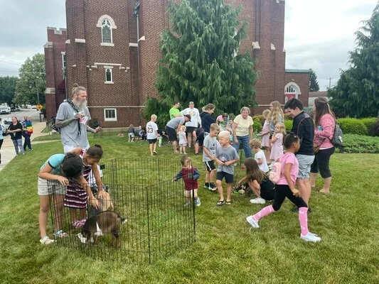 Children and adults gathered outdoors near a brick building with a large tree, interacting with animals inside a small pen, on a grassy area.