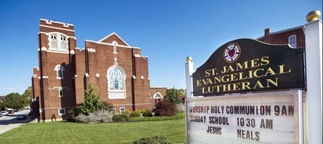 The church building has a brick exterior with pointed towers and stained glass windows. There is a sign in the foreground that reads 'St. James Evangelical Lutheran' and advertises worship services.