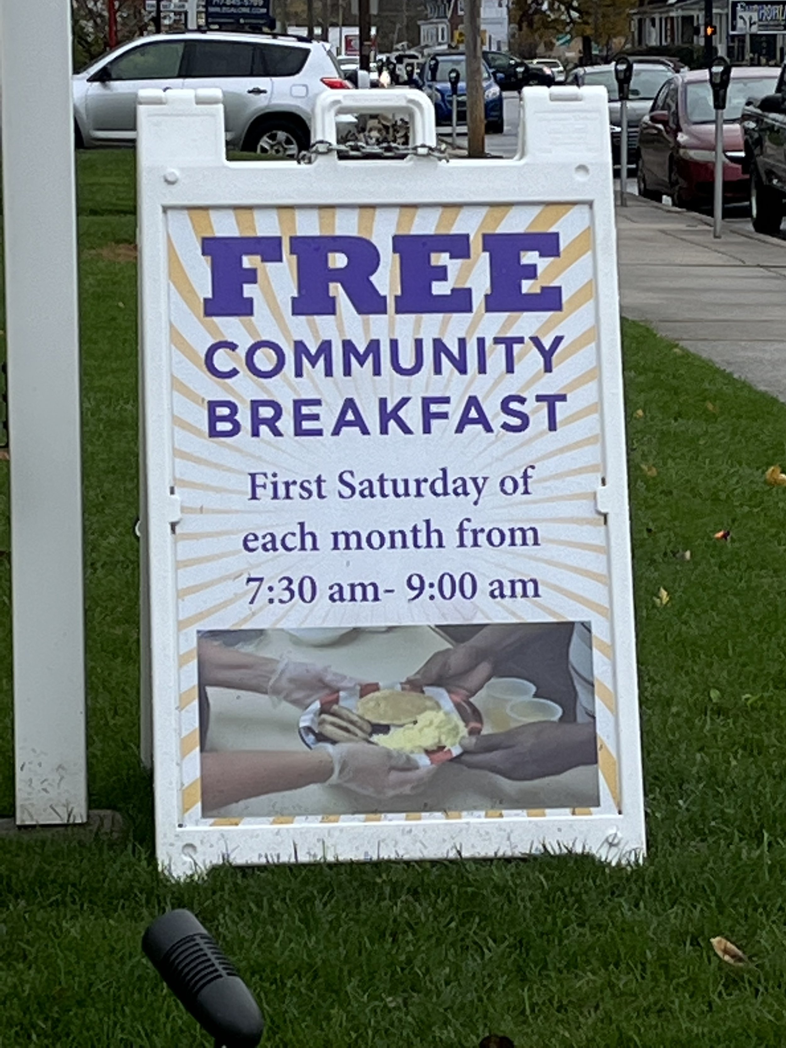 A sidewalk sign advertising a free community breakfast on the first Saturday of each month from 7:30 am to 9:00 am, with a photo of a person holding a plate of breakfast food at the bottom.