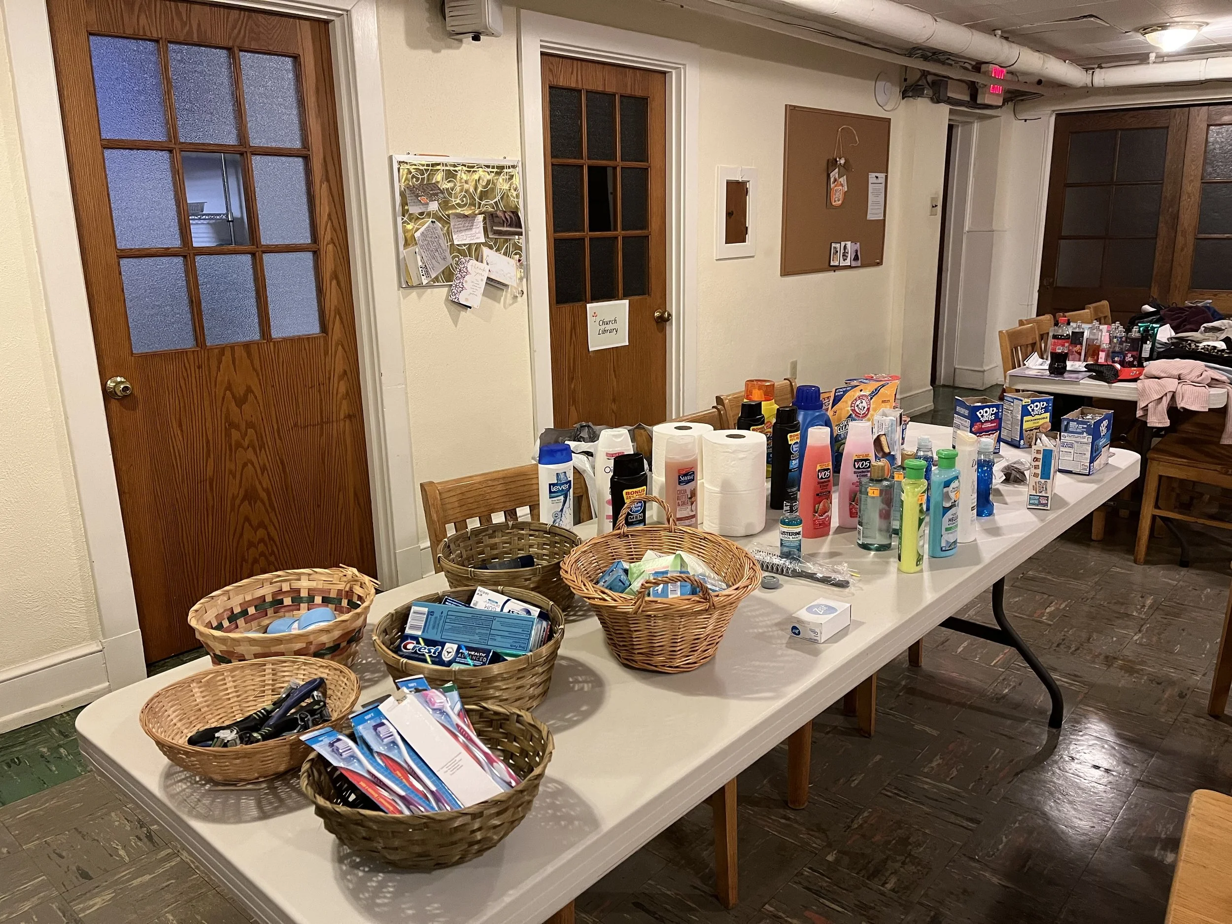 A room with several tables filled with various items, mainly toiletries and personal care products like toothpaste, toothbrushes, and deodorants. Several baskets of miscellaneous items are on the tables, and there are two closed wooden doors on the wall, one with frosted glass panels and a sign reading 'Church Library' hanging on the door handle. There's a bulletin board and various notes and papers pinned on the wall. The floor is dark wood, and part of the ceiling exposes pipes and a mounted speaker.