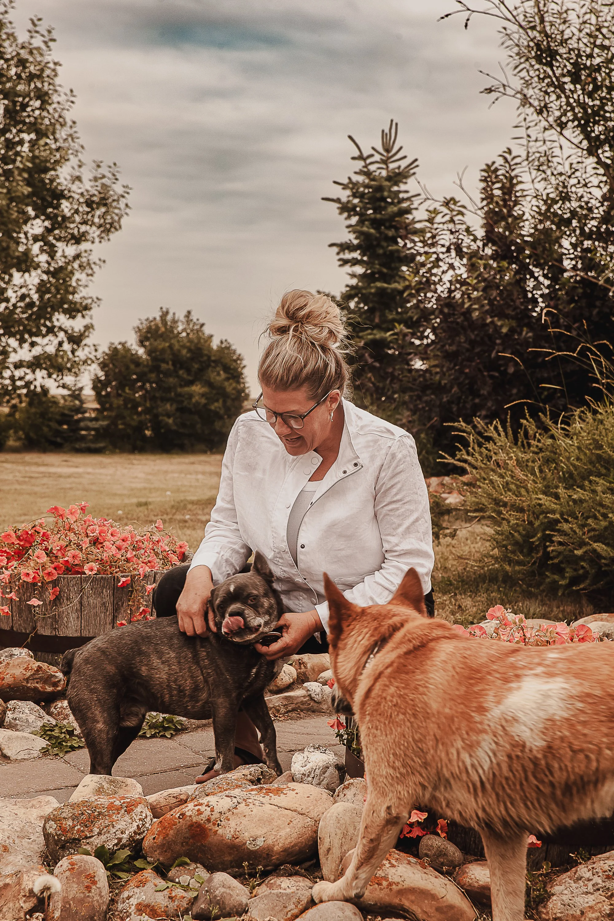 A woman with blonde hair, glasses, and a white jacket interacts with two dogs outdoors on a stone pathway, surrounded by flowering plants, trees, and a cloudy sky.