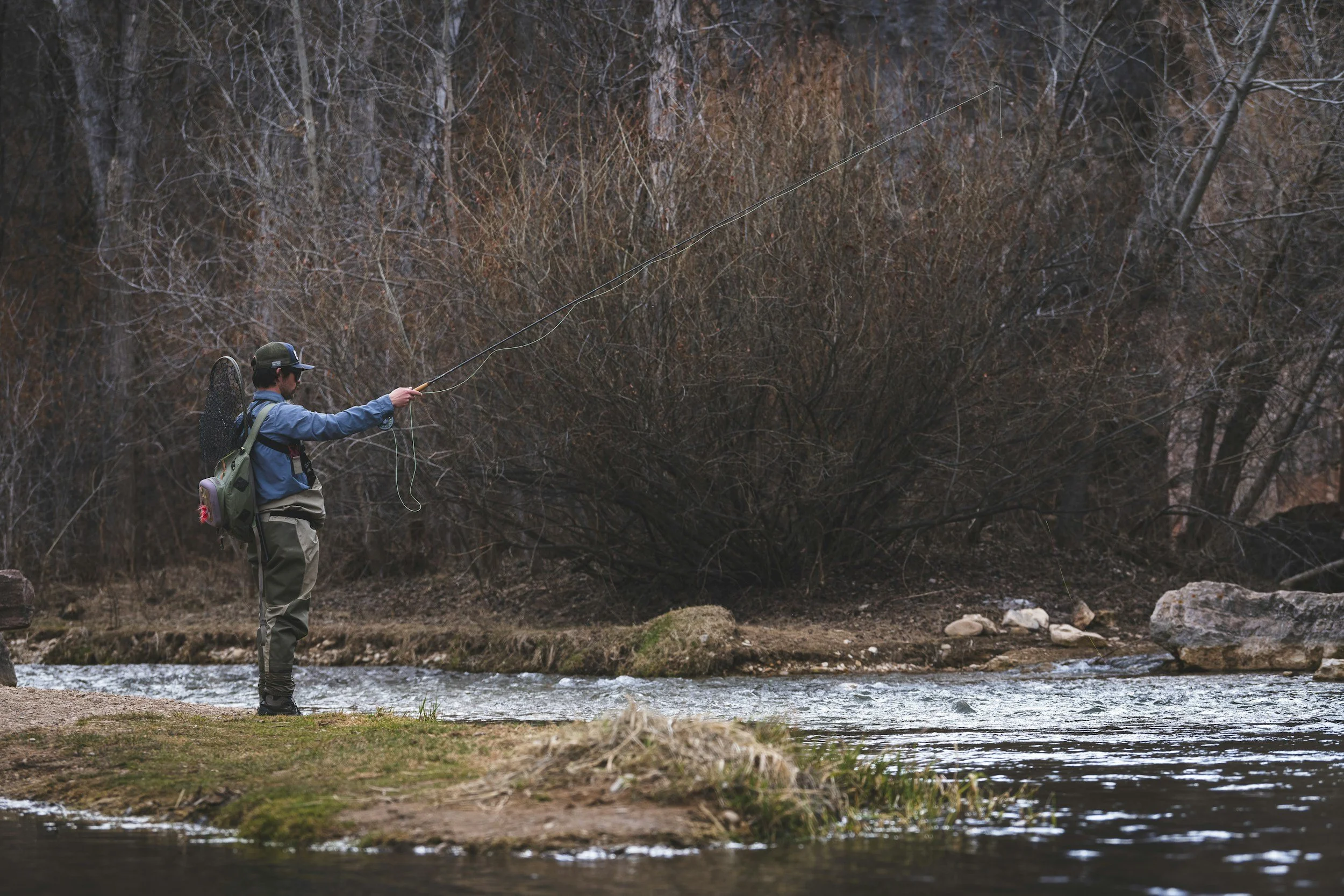 A man fishing in a river surrounded by trees with barren branches.